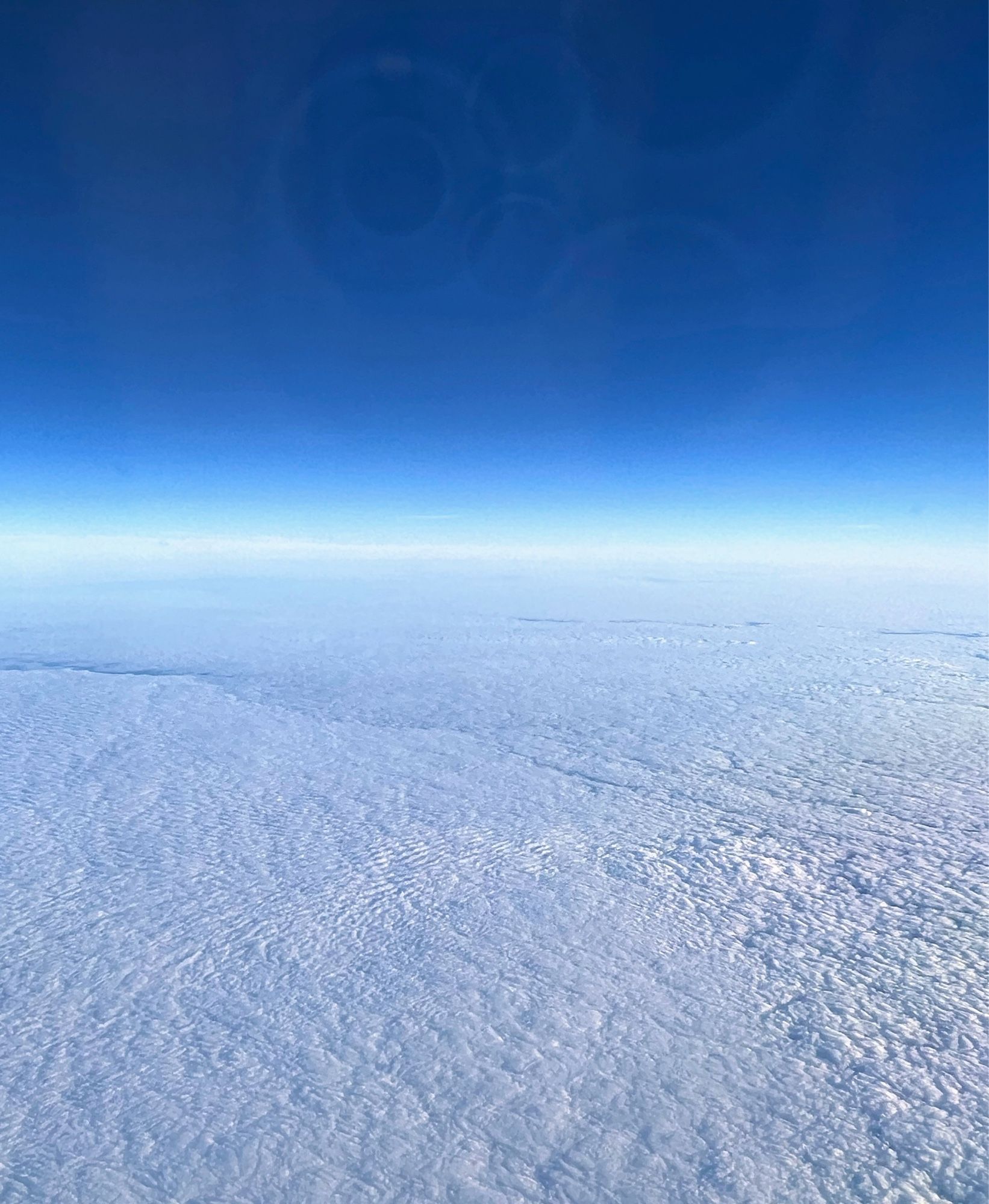 A blanket of clouds, from the perspective of an airplane window, stretches to meet a bright horizon and a clear deep sky. Photo by Karin Hedetniemi.