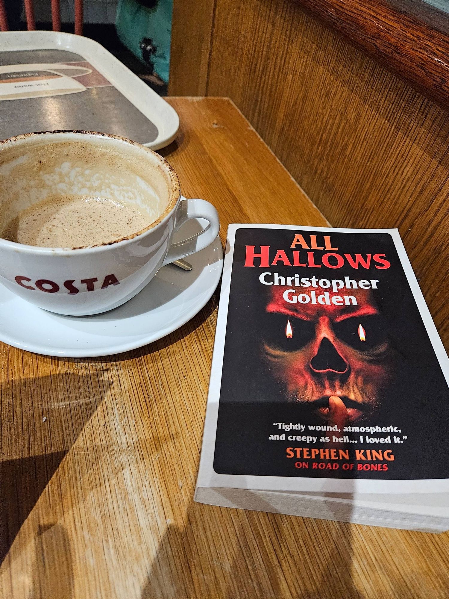 Coffee mug, half full, next to a book titled 'All Hallows' by Christopher Golden. The cover shows red writing on a black background and a skull with a finger to its mouth in a shushing gesture. Candles blaze in the skull's eyes.