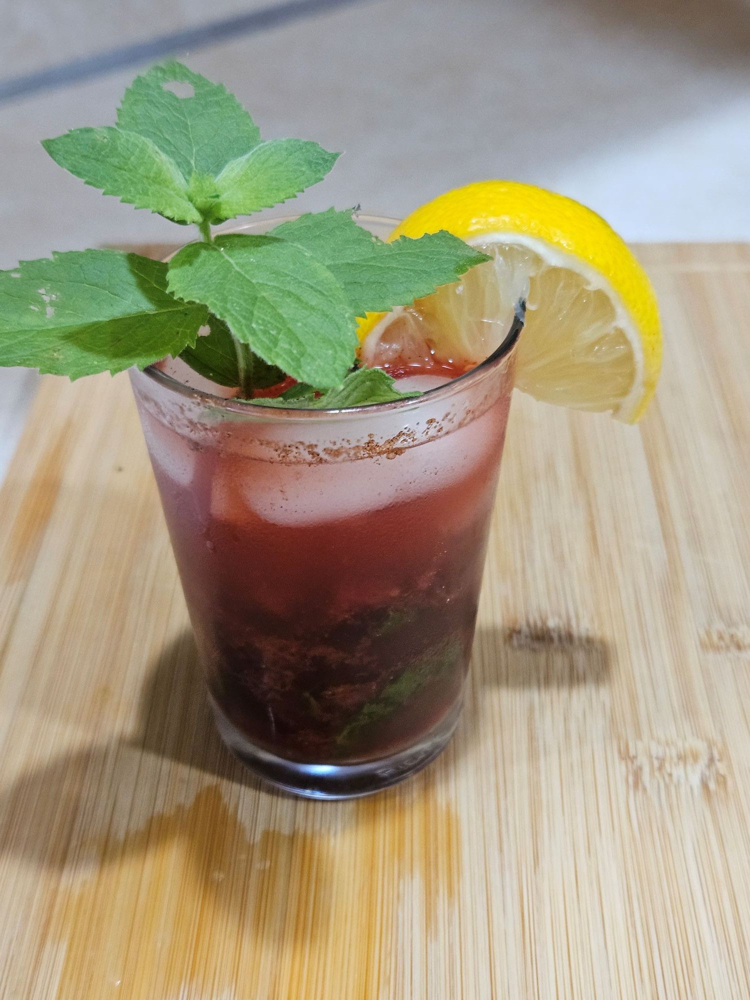 red-toned drink in a glass with ice. garnished with bright green mint leaves and a lemon slice