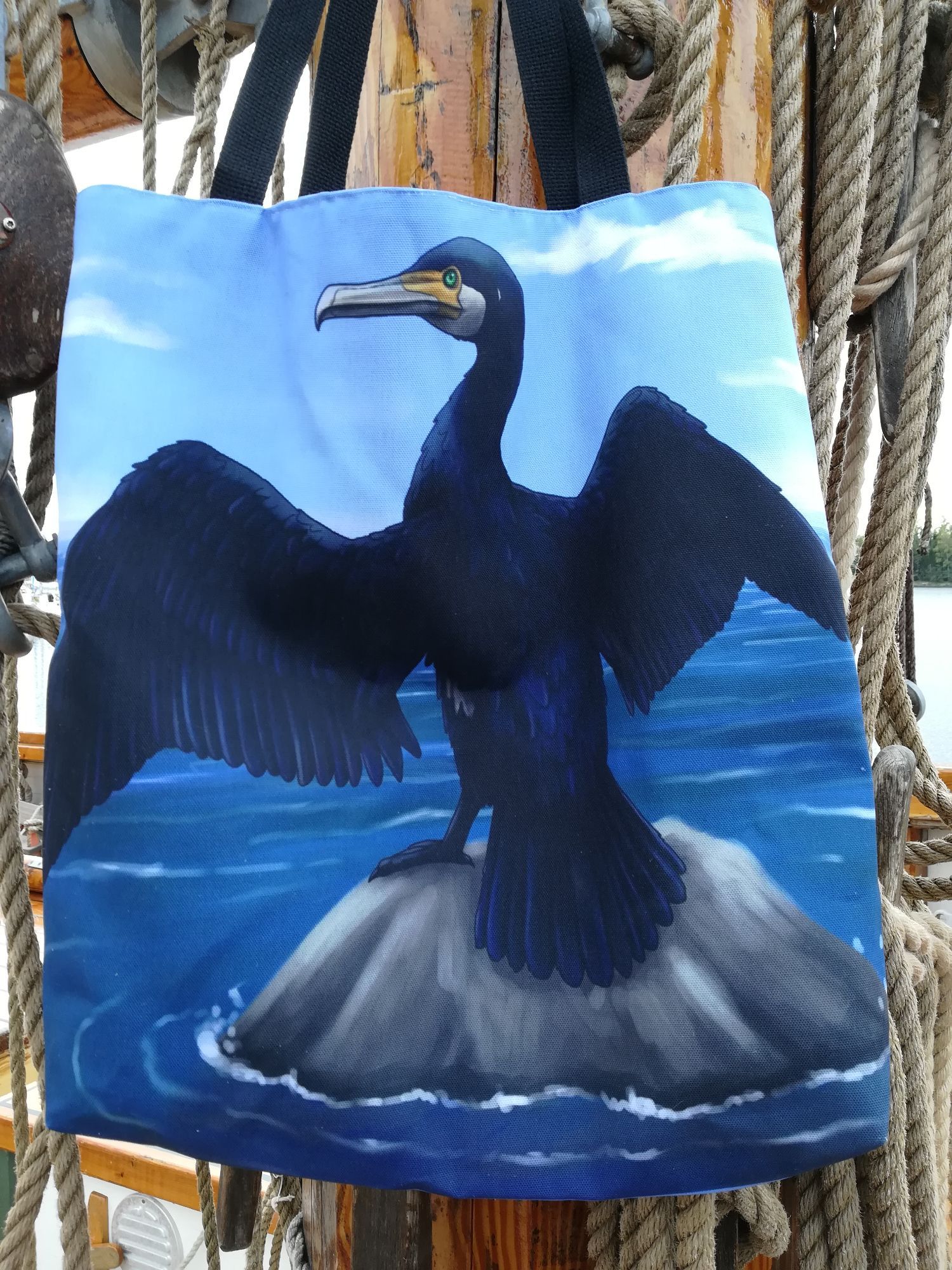 A photo of a tote bag with an illustration of a cormorant on it. The bird is standing on a rock in the middle of the sea and spreading its wings wide. The bag is hanging from the mast of a ship.