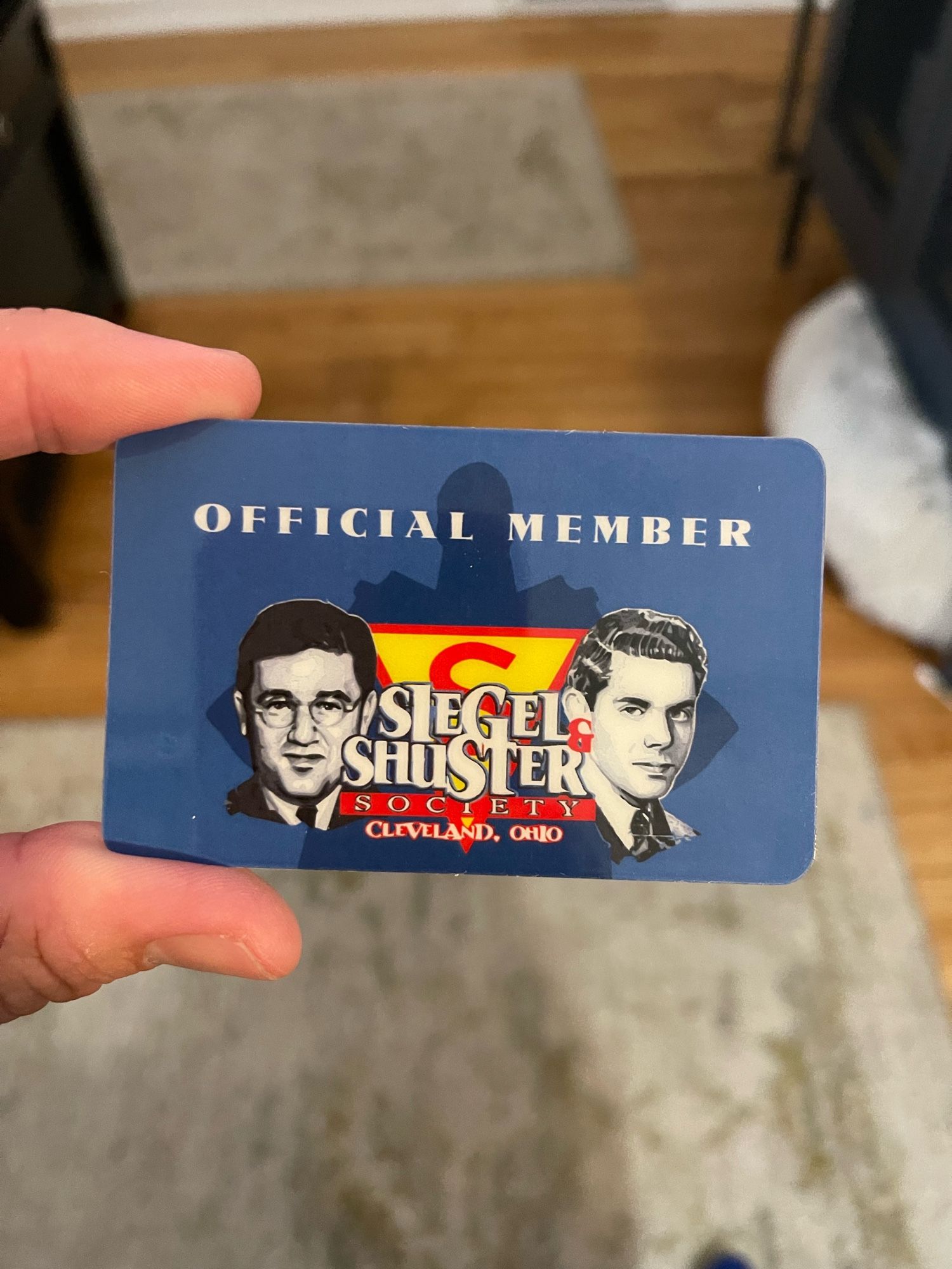 A membership card for the Siegel & Shuster Society, featuring images of two men and the text "OFFICIAL MEMBER" along with "CLEVELAND, OHIO." The background is blue with a logo featuring the letters "S" and "&