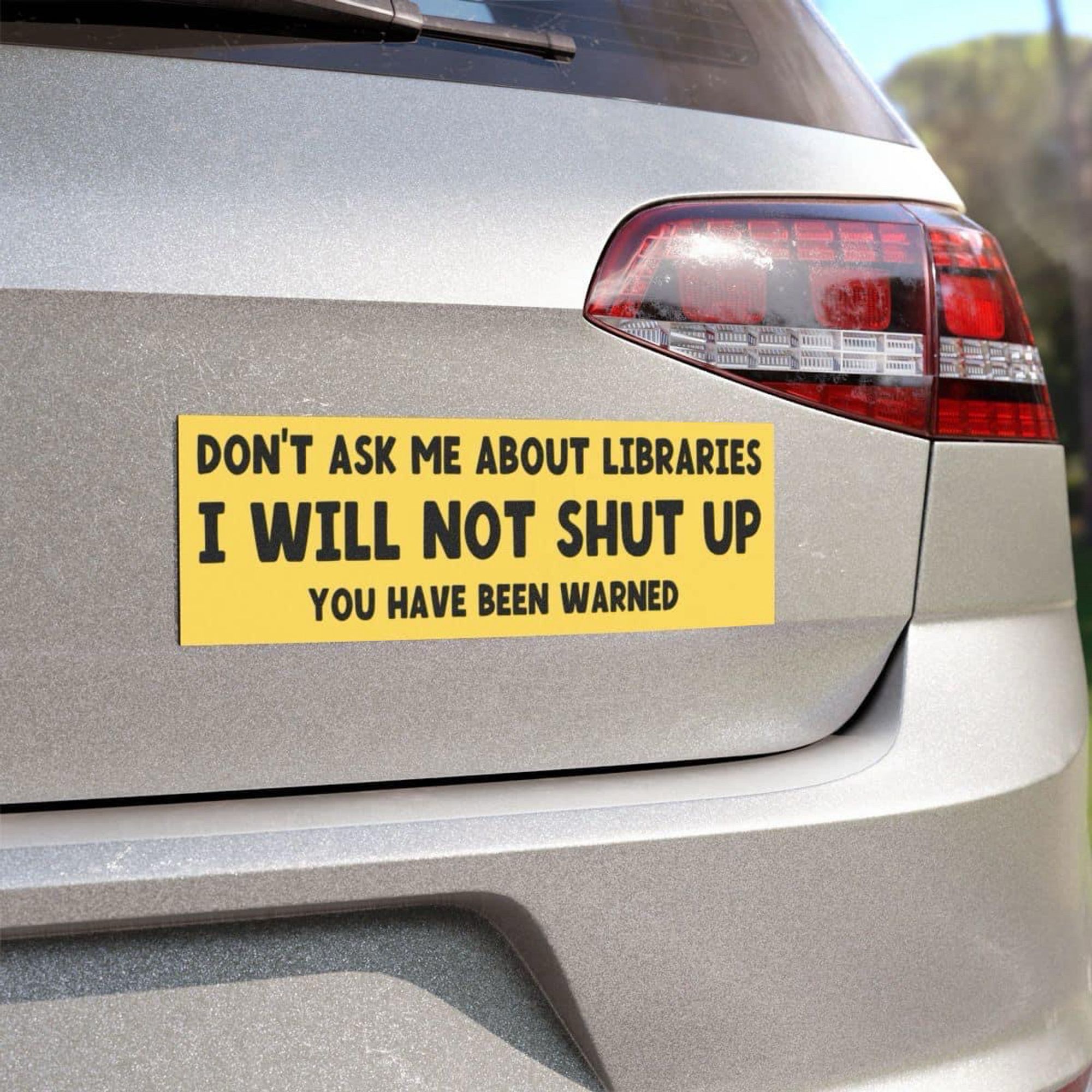 A yellow bumper sticker on the back of a silver vehicle reads: "DON'T ASK ME ABOUT LIBRARIES I WILL NOT SHUT UP YOU HAVE BEEN WARNED."