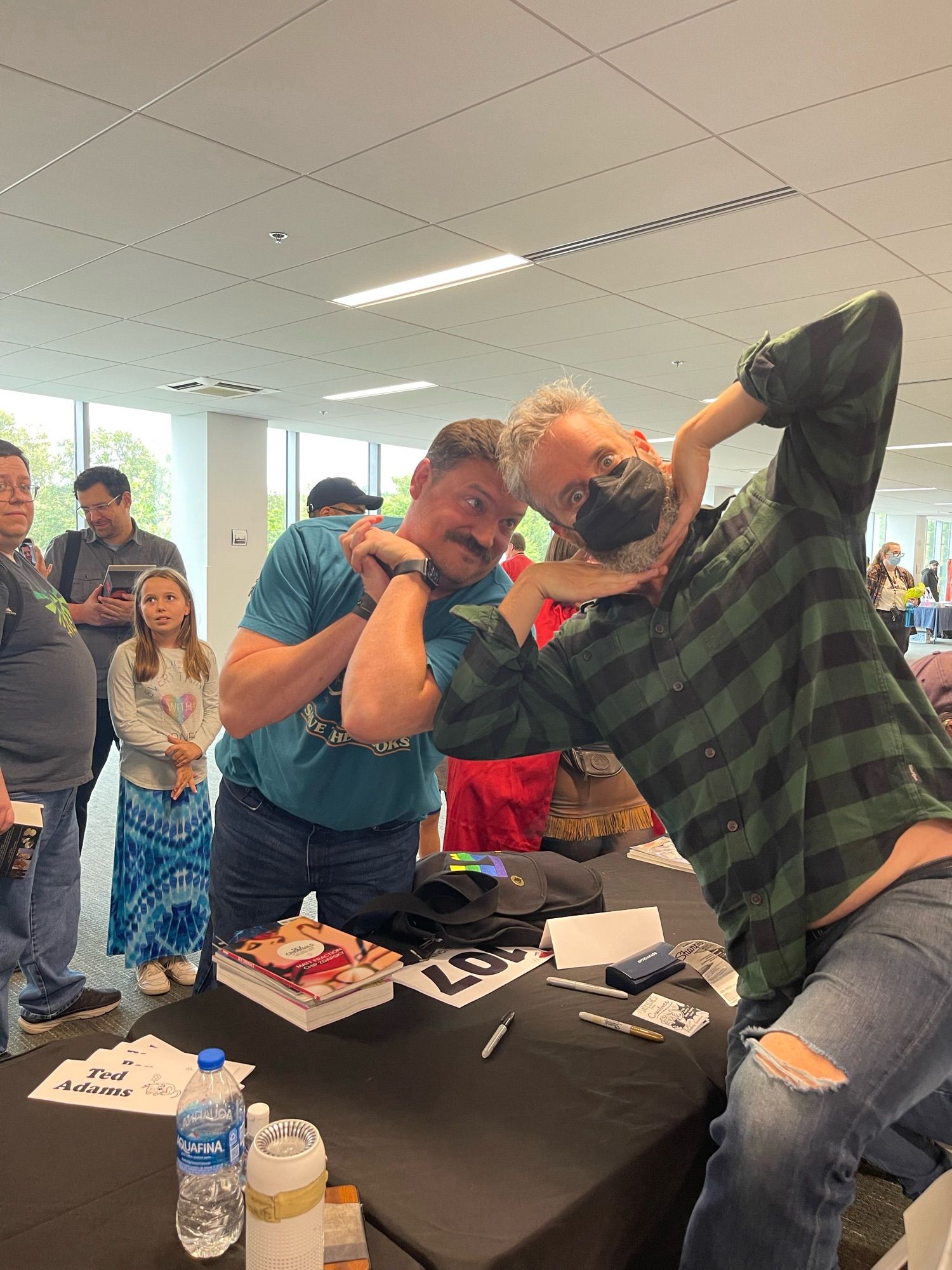 Two men strike humorous poses at a table, surrounded by fans. One man wears a black mask and a green plaid shirt, while the other is in a blue t-shirt. A child stands nearby, and various books and items are on the table.