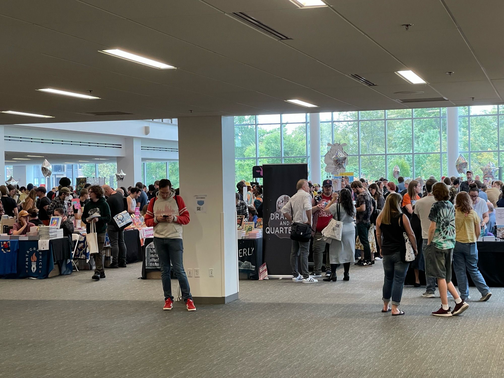 A bustling indoor event with many people gathered around tables displaying books and merchandise. The setting features large windows allowing natural light, and groups of attendees are engaged in conversations and browsing items. Some individuals are actively using their phones.