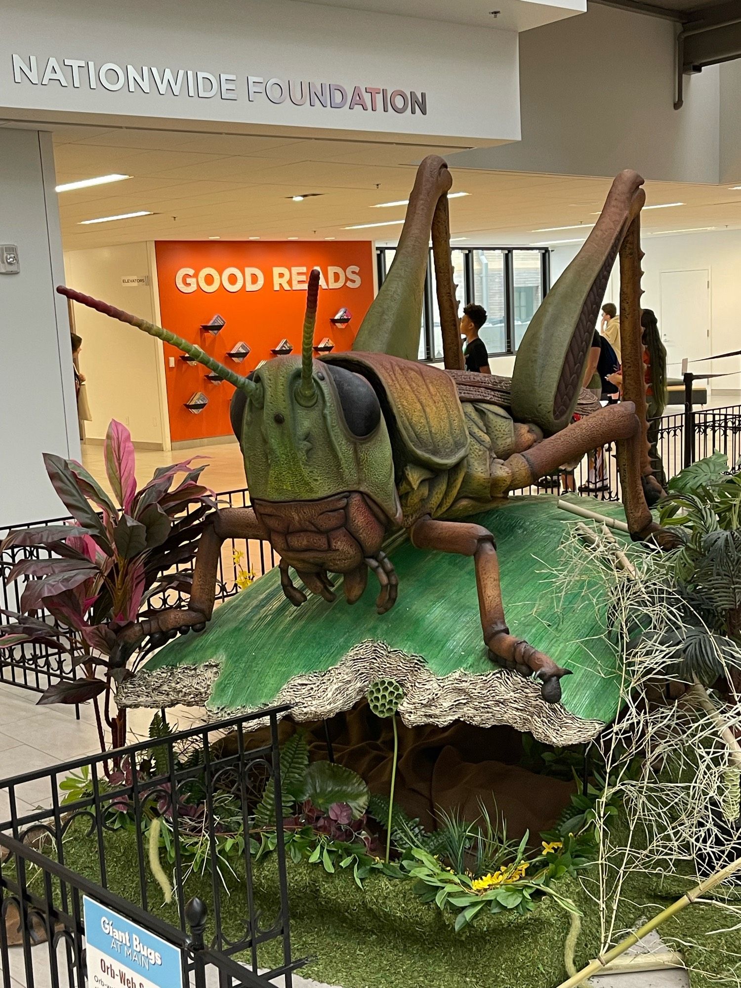 A large, realistic sculpture of a grasshopper sits on a green leafy structure, surrounded by artificial plants. In the background, the text "Nationwide Foundation" and "Good Reads" is visible on the walls of a bright indoor space.