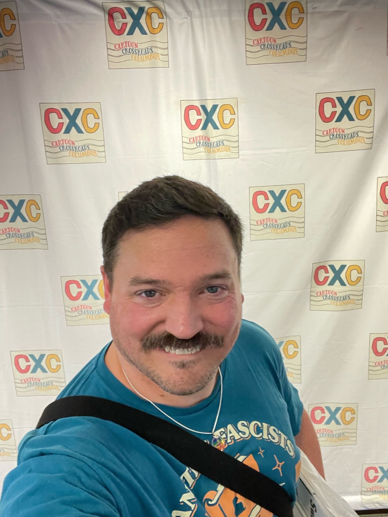 A smiling man with a mustache poses for a selfie in front of a backdrop featuring the logo for "CXC: Cartoon Crossroads Columbus." He wears a blue shirt and has a bag slung over his shoulder.