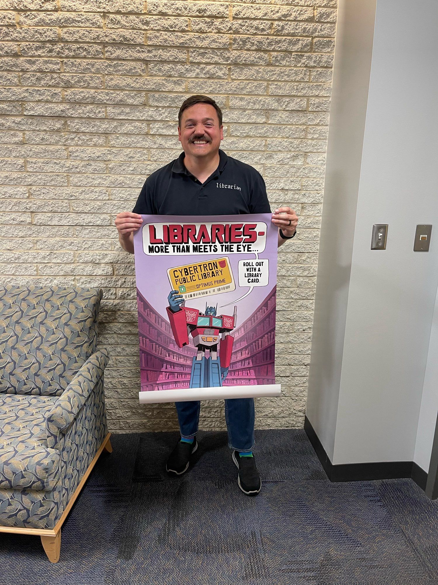 A man with a mustache is holding a poster that promotes libraries, featuring a cartoon character resembling Optimus Prime amidst shelves of books. The poster reads, "Libraries: More Than Meets the Eye..." and mentions "Cybertron Public Library." The background