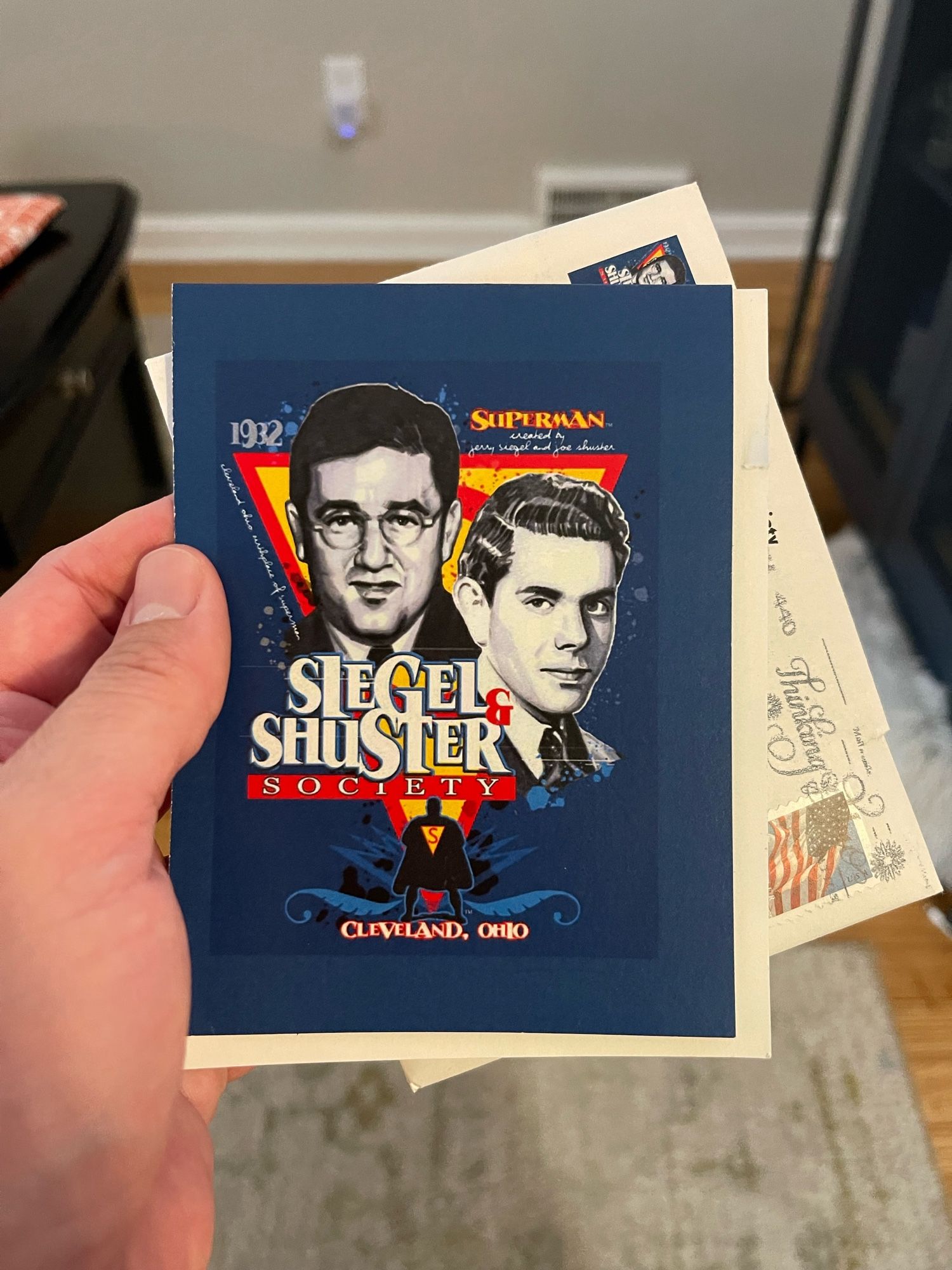 The image features a hand holding a card that celebrates the Siegel & Shuster Society, highlighting the creators of Superman. The card includes illustrations of Jerry Siegel and Joe Shuster, with the year "1932" prominently displayed. A folded envelope