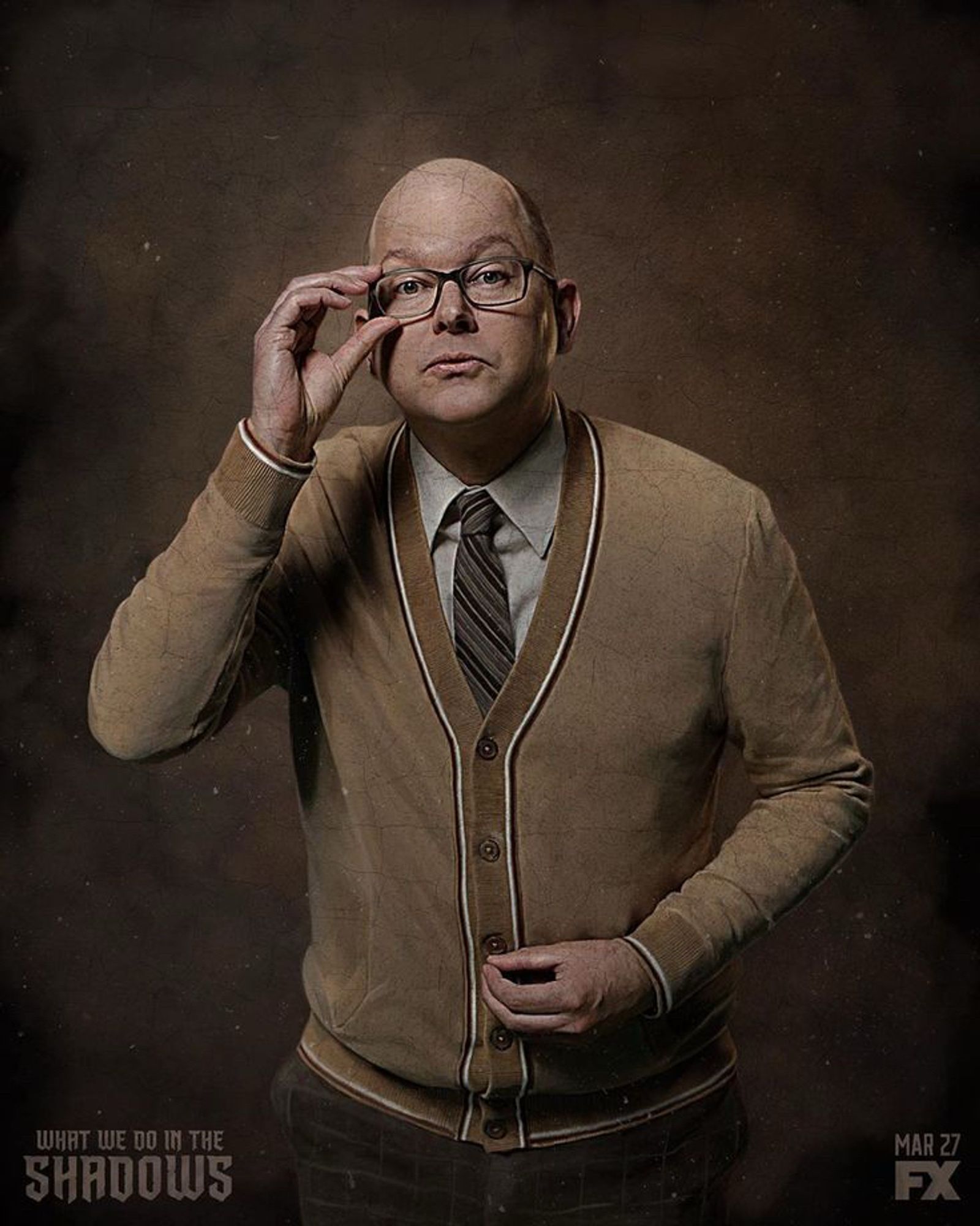 A promotional photo of Colin Robinson from What We Do In The Shadows. He is wearing a brown sweater vest and holding is rimmed glasses while looking into the camera. 