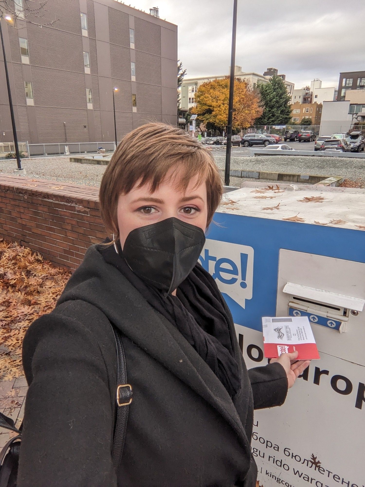 A picture of a woman with short brownish red hair wearing a black strapless kn95 standing in front of a voting Drop Box about to drop her ballot in it