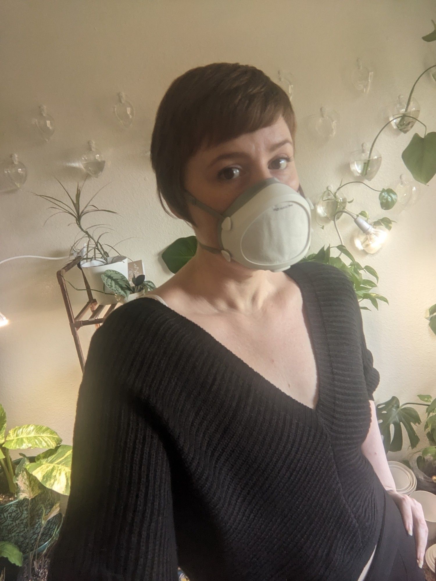 A picture of a woman with short brownish red hair and brown eyes wearing a full mask and a black sweater and black trouser behind her are a lot of plants