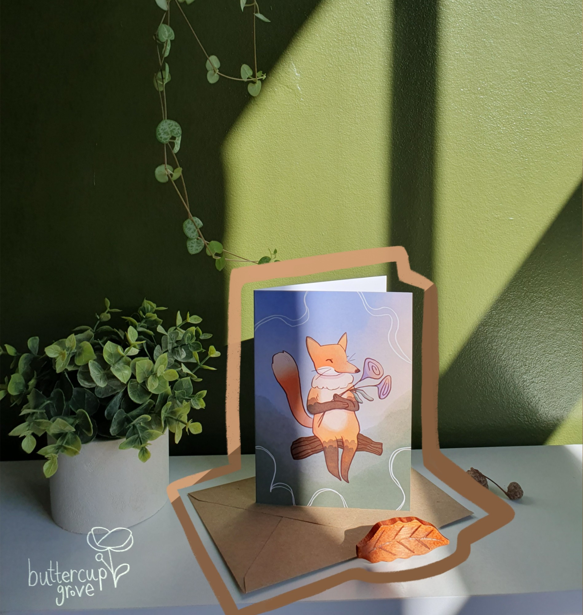 photo of a green wall and a white shelf infront, in a sunny room with a plant and a greetings card sat on the shelf. the card is art of a happy orange fox sat on a log holding 2 purple flowers with blue and green watercolour textures behind. drawn around the shape of the card and envelope is an orange and brown coffee coloured line. in the bottom left corner is a watermark of buttercup grove and a simple buttercup logo.