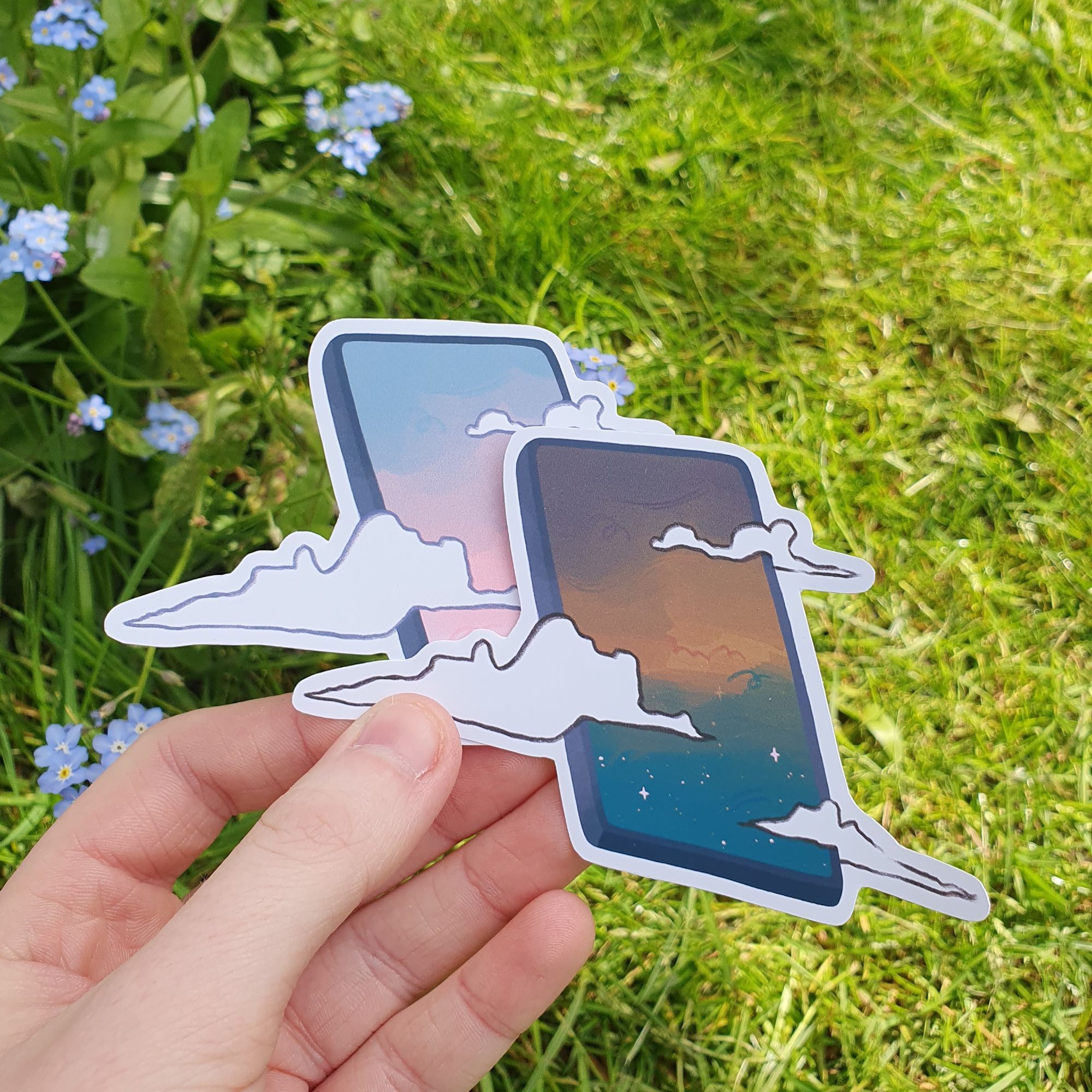 square photograph of a white hand holding two cut out stickers, the art on them is of mobile phones with colourful skies inside and white clouds passing over the outside of the phone. in the background is a sunny grassy environment and small blue flowers (forget me nots).