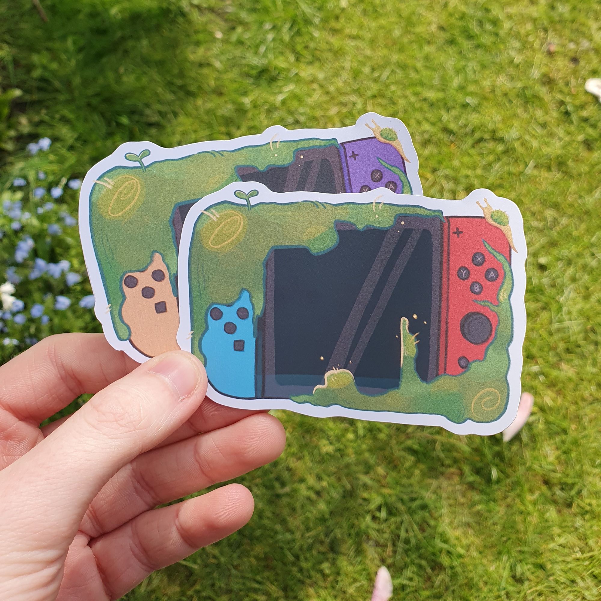 square photograph of a white hand holding two cut out stickers, the art on them is of nintendo switch consoles in different colours, and they are partially covered in green moss. in the background is a sunny grassy environment.