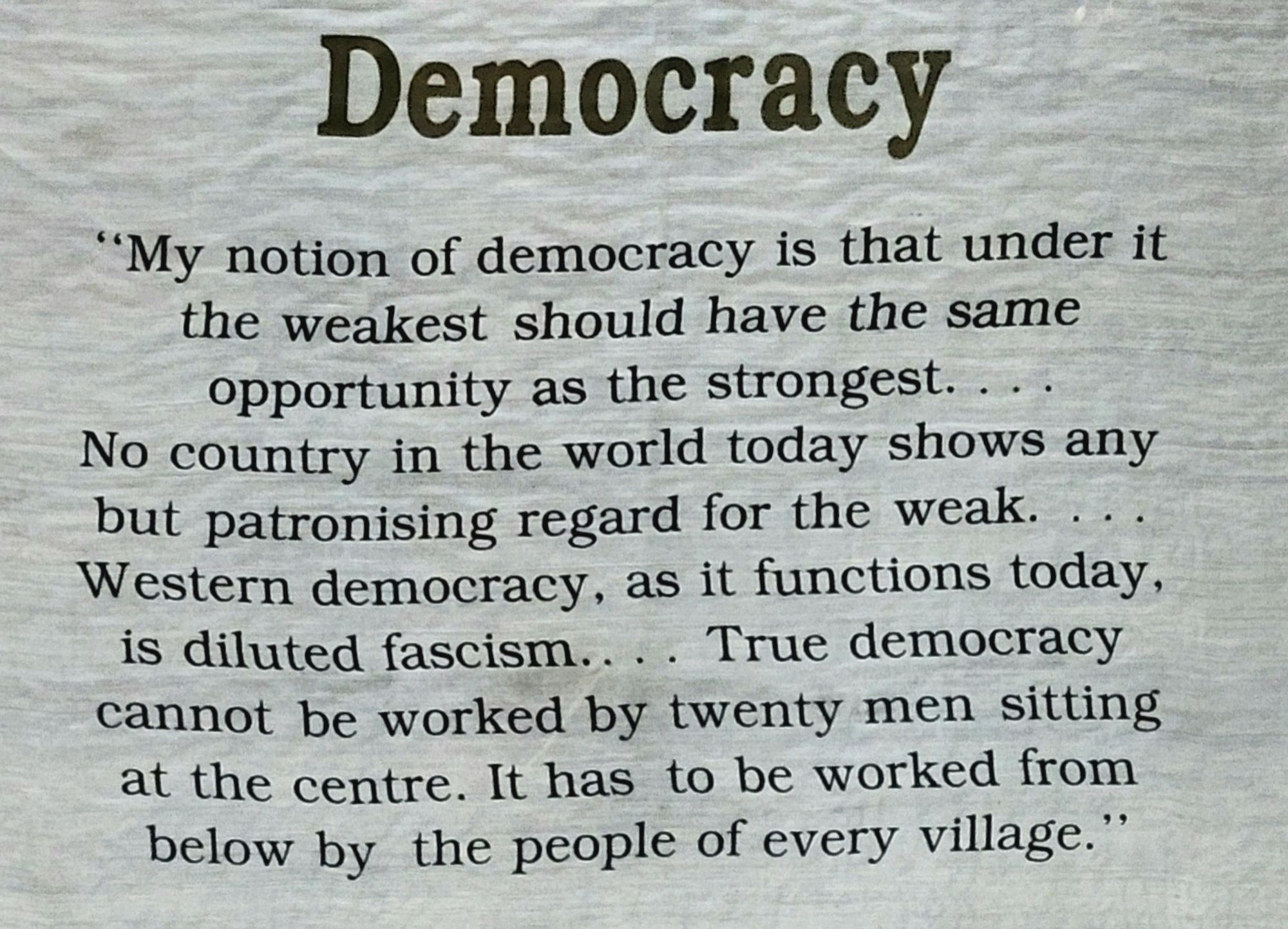 A white poster with black text in English:

Democracy

"My notion of democracy is that under it
the weakest should have the same
opportunity as the strongest. ...
No country in the world today shows any
but patronising regard for the weak. ...
Western democracy, as it functions today,
is diluted fascism. ... True democracy
cannot be worked by twenty men sitting
at the centre. It has to be worked from
below by the people of every village."