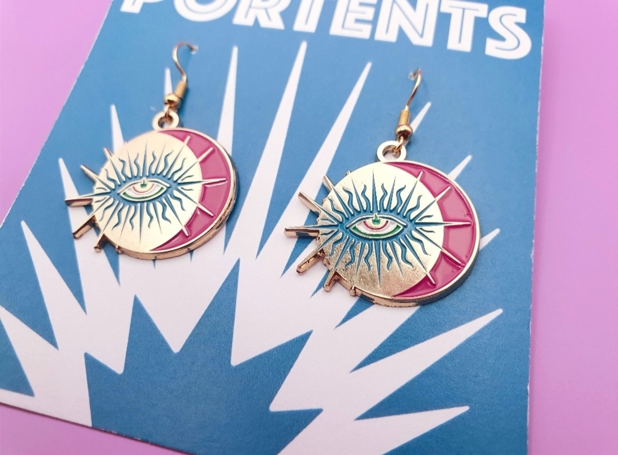 A photo of a pair of earrings in enamel with gold plated metal. They are circular and feature a pink crescent moon, and an eye in blue rays. Gold spikes stick out the left side of each circle. There is a flame in the pupil of the eye.