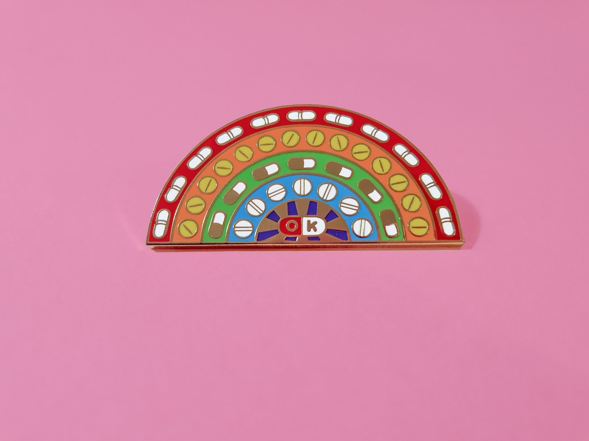 A photo of a rainbow shaped enamel pin with different shaped pills in each of its 4 arcs. In the central semicircle is a half red half white pills with the letters O K on it.