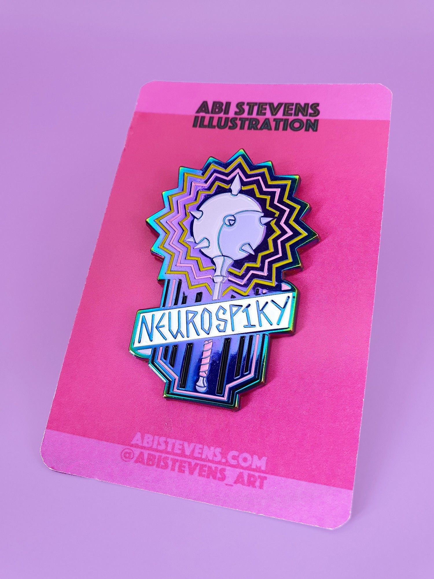 A photo of a rainbow plated enamel pin featuring a lilac mace with spiky rings of pink and yellow around the head and a white banner across the handle reading: Neurospiky.