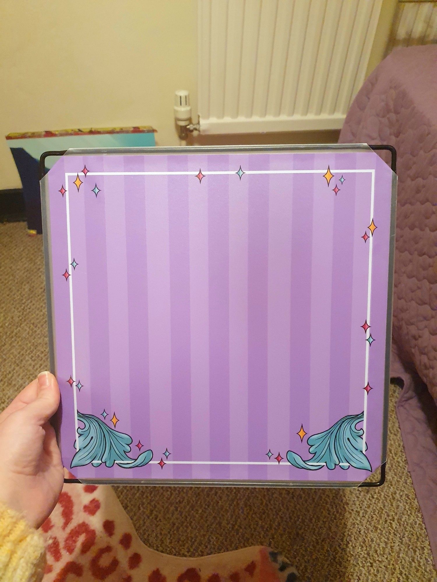 A photo of my hand holding a plastic square from the grid box shelving kits a lot of artists use for building convention displays. This one has a vinyl square stuck on it in the lilac stripes of my branding with little leafy details in the bottom corners and some sparkles dotted around the edges.