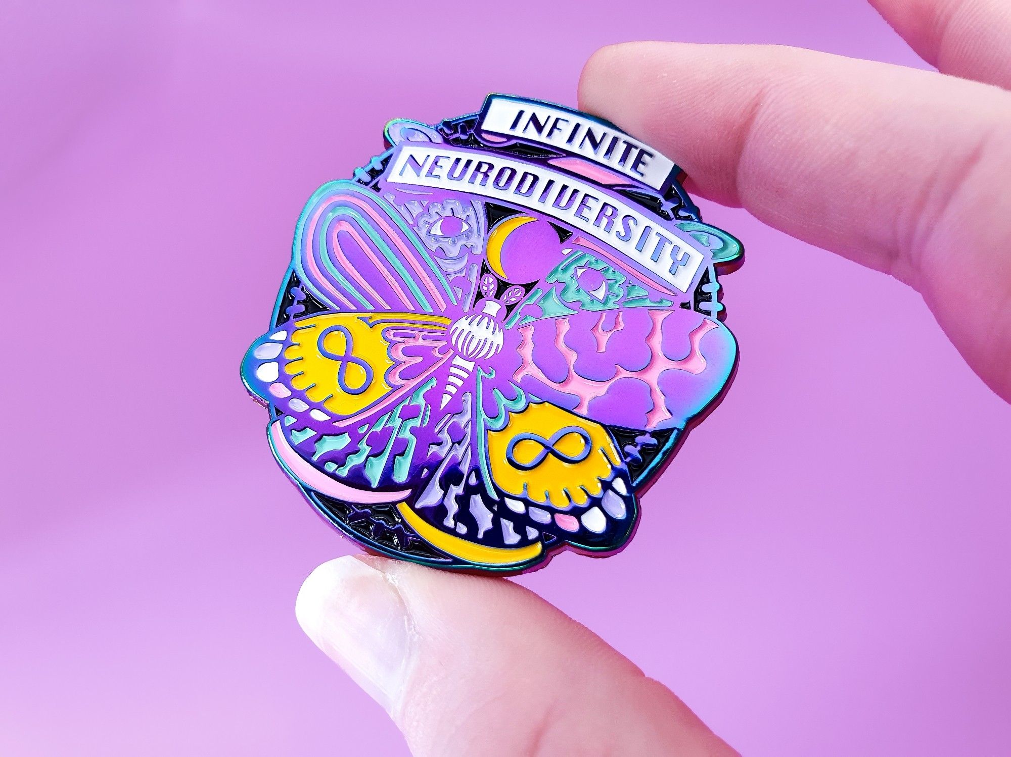 A photo of a rainbow plated enamel pin featuring a moth with 8 different wings and a banner reading: infinite neurodiversity.