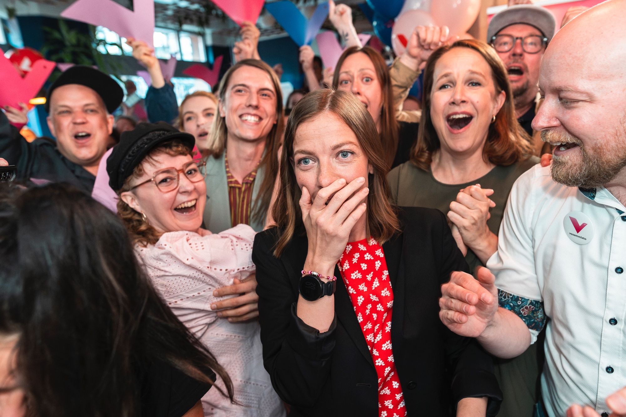 A group of people are celebrating. The focal point is a woman in the center with long hair, wearing a black blazer over a red dress with white floral patterns. She is covering her mouth with her hand, her eyes wide with excitement and surprise. Around her, other people are cheering, laughing, and smiling.