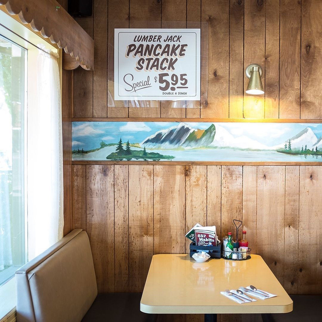 small white vinyl booths with an open window behind it, a cream colored table, wood shiplap walls with a border of a painting with a lake/mountains/trees motif and a sign that says “Lumber Jack Pancake Stack - Special - $5.95”