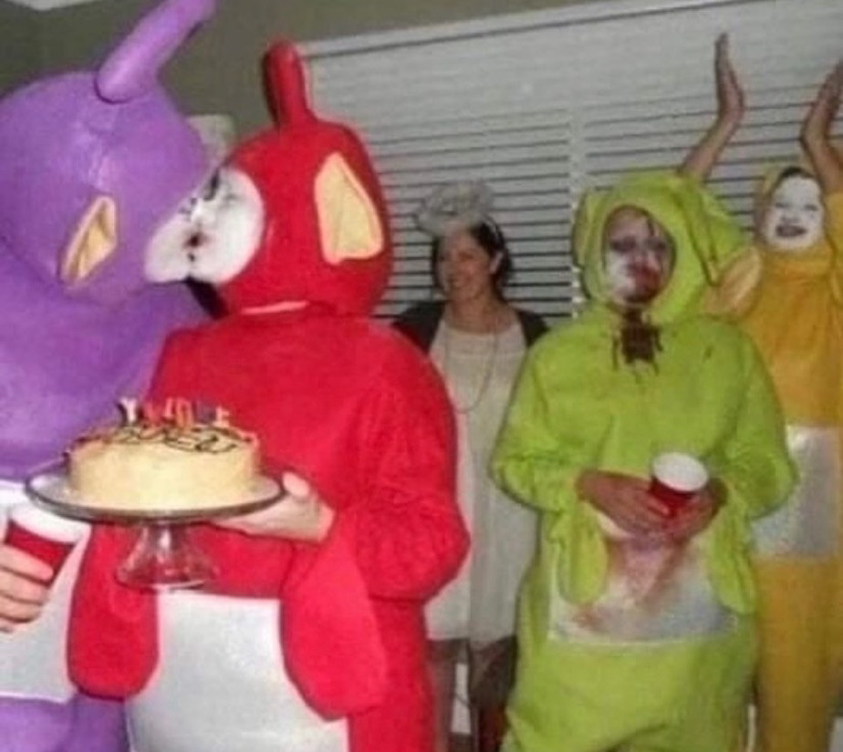 a party where almost everyone is dressed like a Teletubby. 2 Teletubbies are holding a cake and kissing. 1 Teletuby has something dark all over it’s front that looks like they puked it up. 1 Teletubby has their face painted like a mime and is clapping. And 1 person is not dressed like a Teletubby, but is dressed like an angel. 