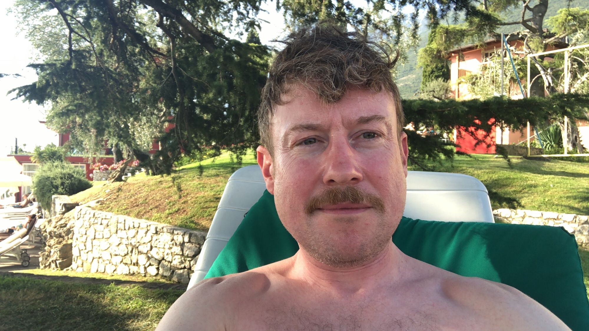 A slightly pink looking white man with reddish brown hair is sat on a sunlounger under a pine tree in am italian garden