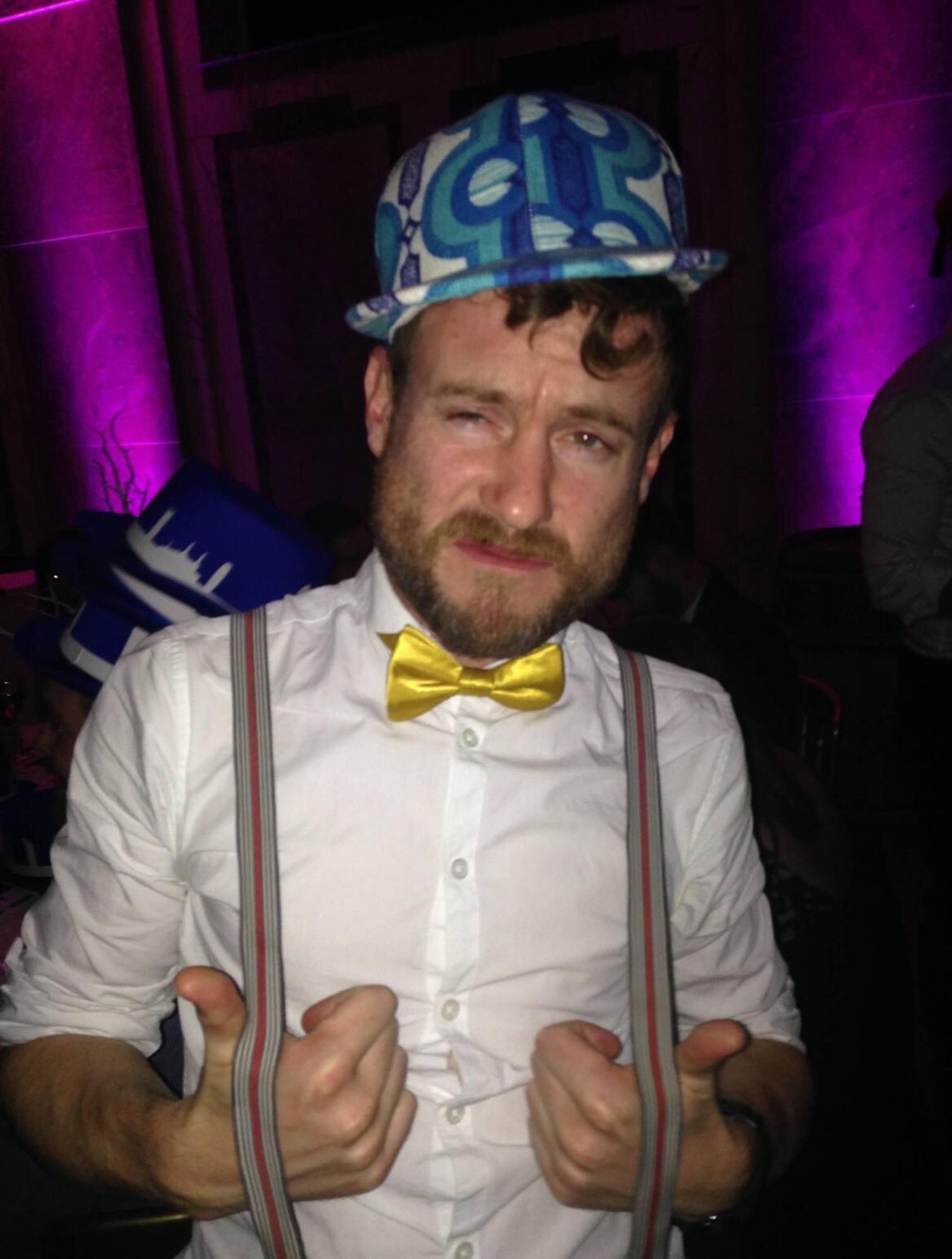 A white mid 30s gay with a short brown beard and moustache has his thumbs in his braces, a white shirt, gold bow tie, and a blue and white 60s patterned baseball hat.