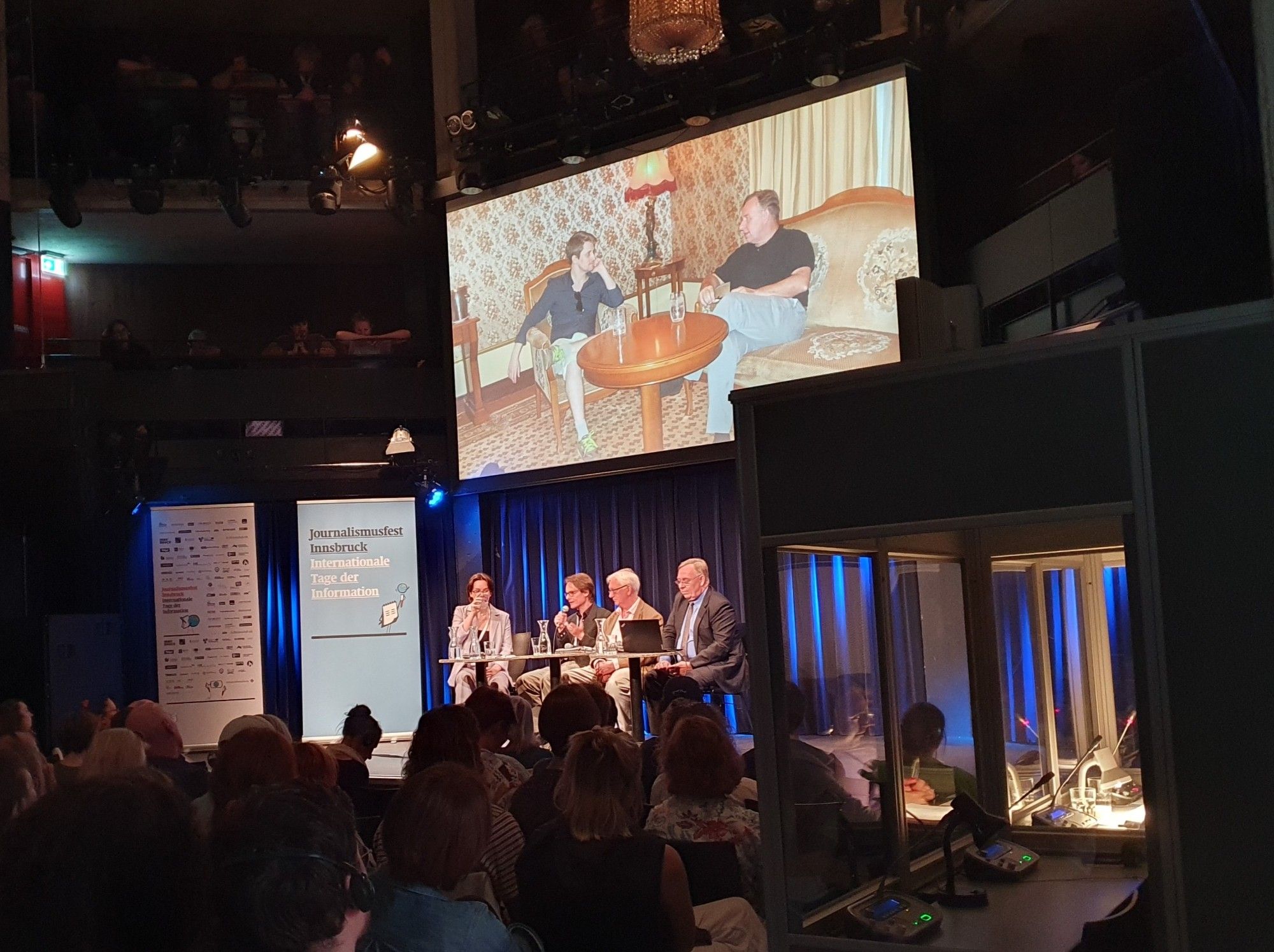 Vier Personen auf einem Podium, darüber ein Bildschirm mit einem Foto und davor Publikum