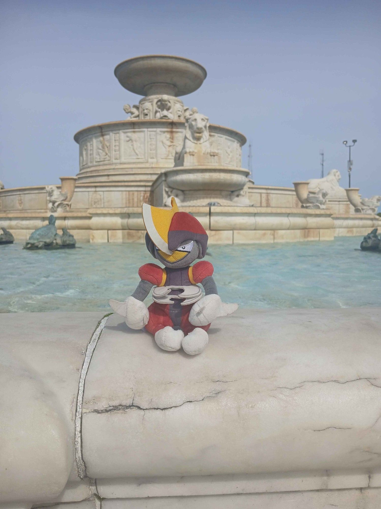 A Bisharp plush sitting on the edge of a fountain carved with lions and turtles. The fountain is not running.