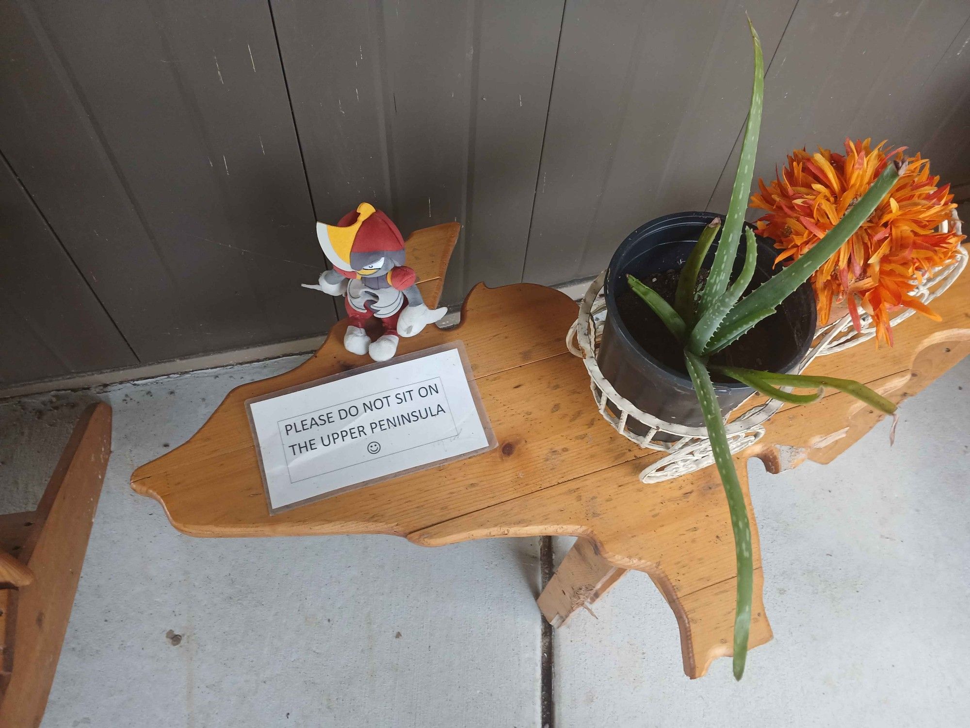 A Bisharp plush on a table shaped like the northern portion of Michigan, called the Upper Peninsula. There are two succulents on the table, as well as a paper that says 'please do not sit on the upper peninsula'