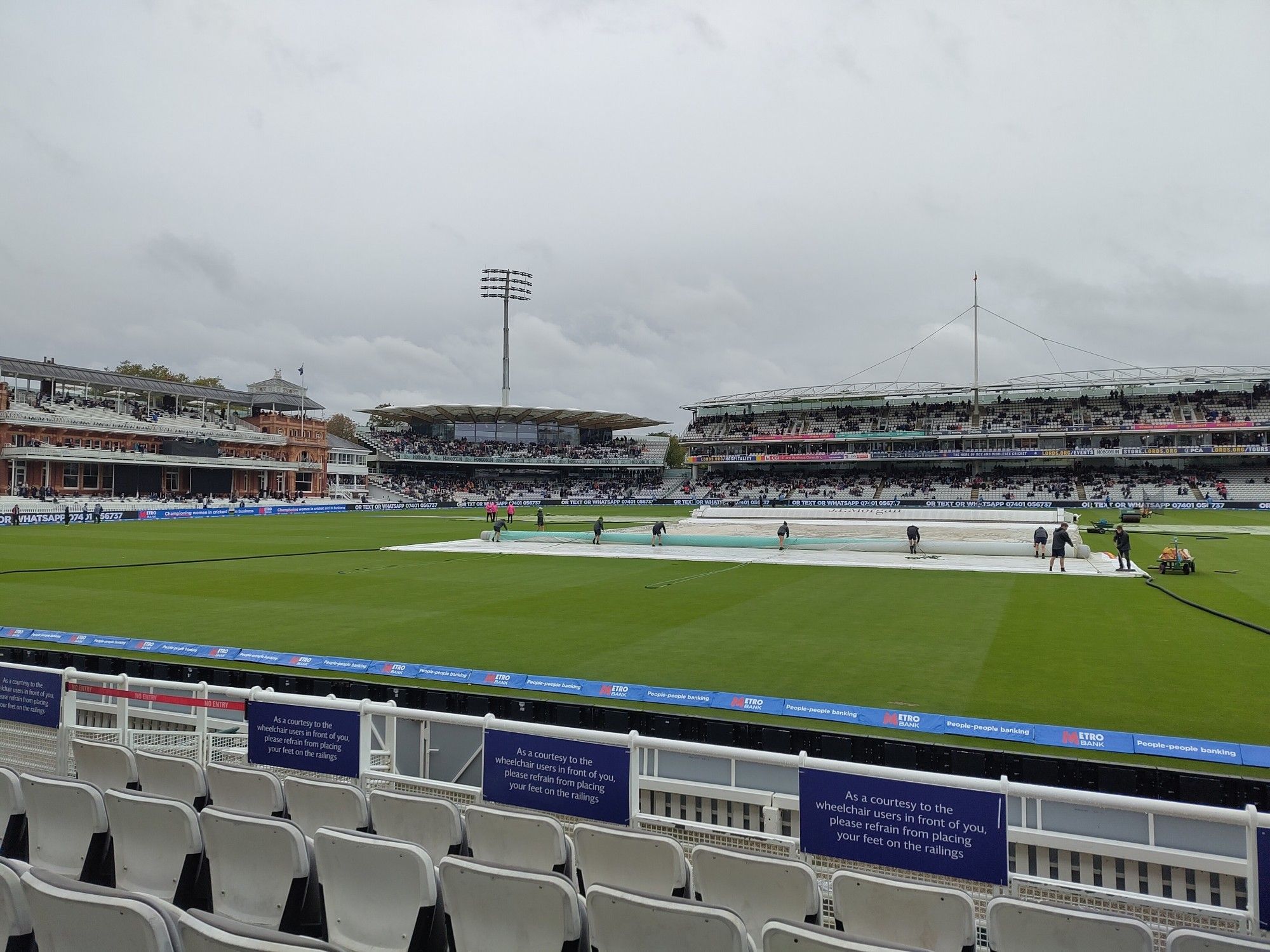 View over the pitch at Lord's. Manky grey sky. Covers are being addressed by groundspeople wearing shorts (as is Traditional) and wielding brooms.