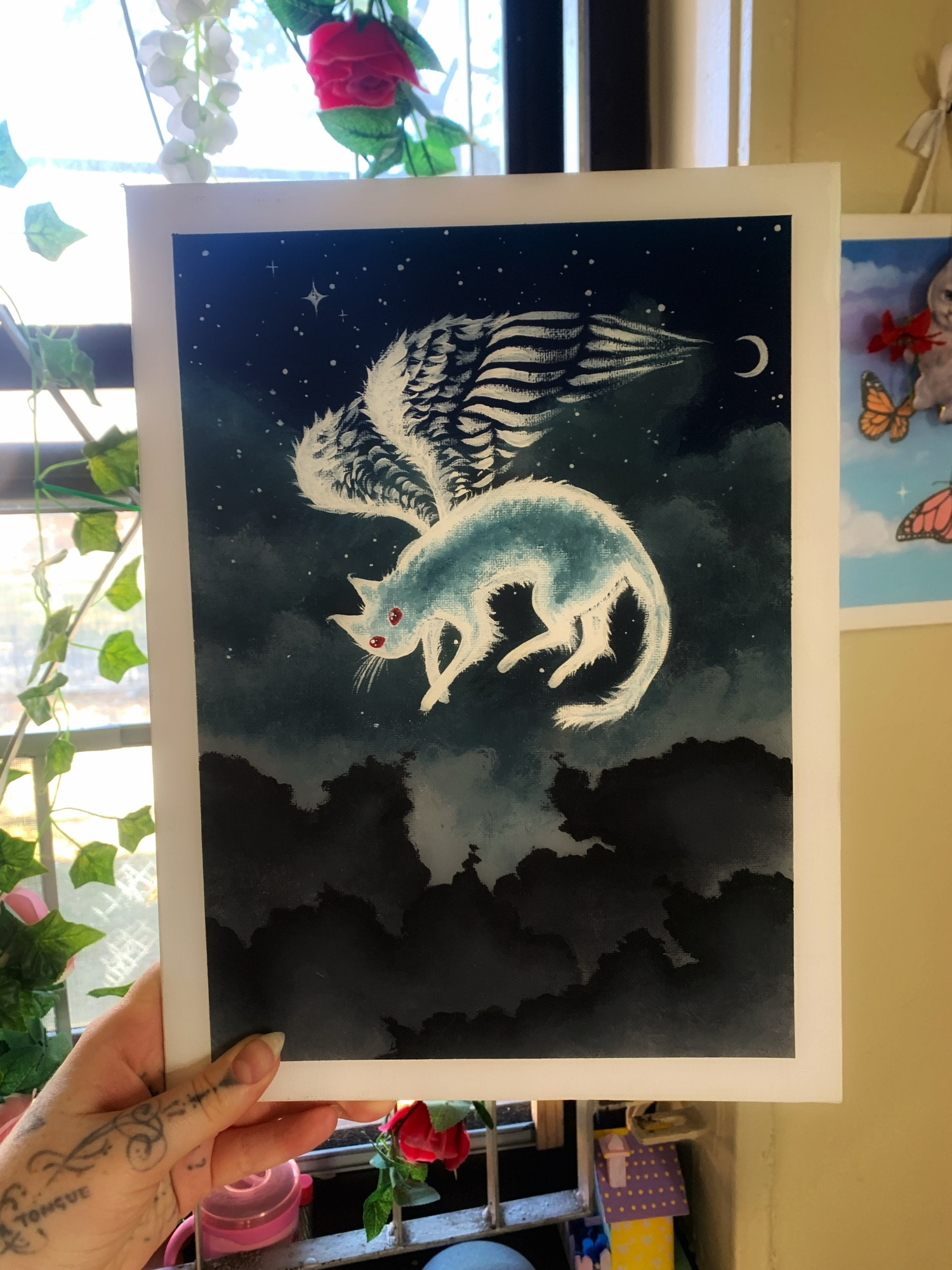 Thea holds a canvas that she painted. On it is a transparent white ghost cat with red eyes and white angel wings. Behind it is the night sky with stars, dark fluffy clouds and a crescent moon. 