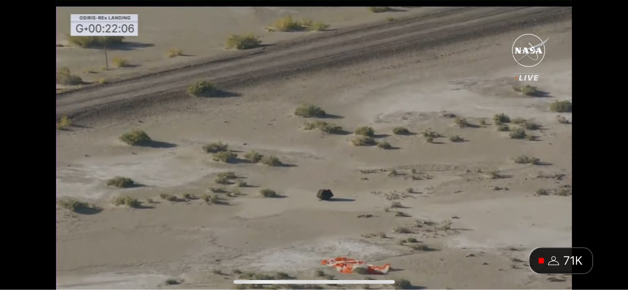 A capsule sitting in the desert with a few shrubs and the orange and white parachute visible