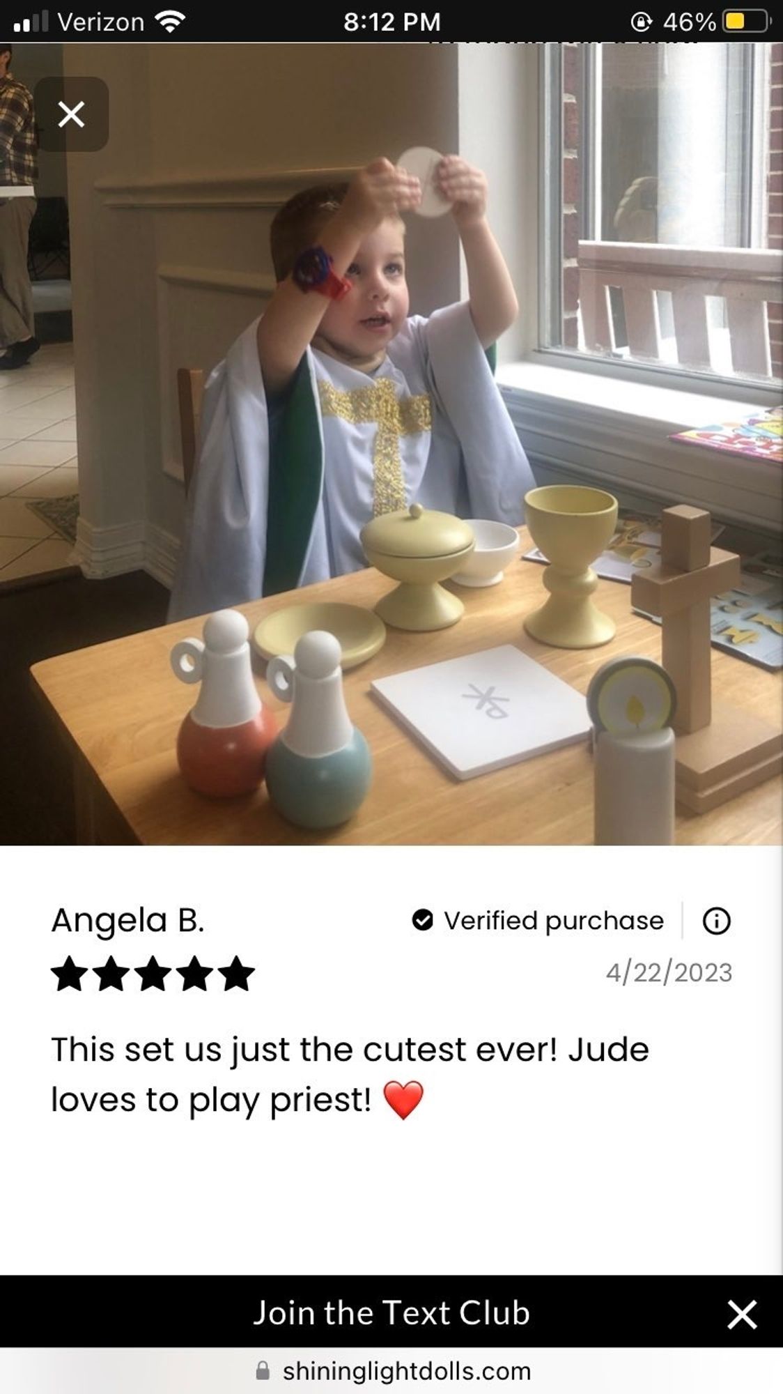 White child holding up circle in front of table with wooden toy goblets, cross, etc, wearing a tiny white alb with a yellow cross