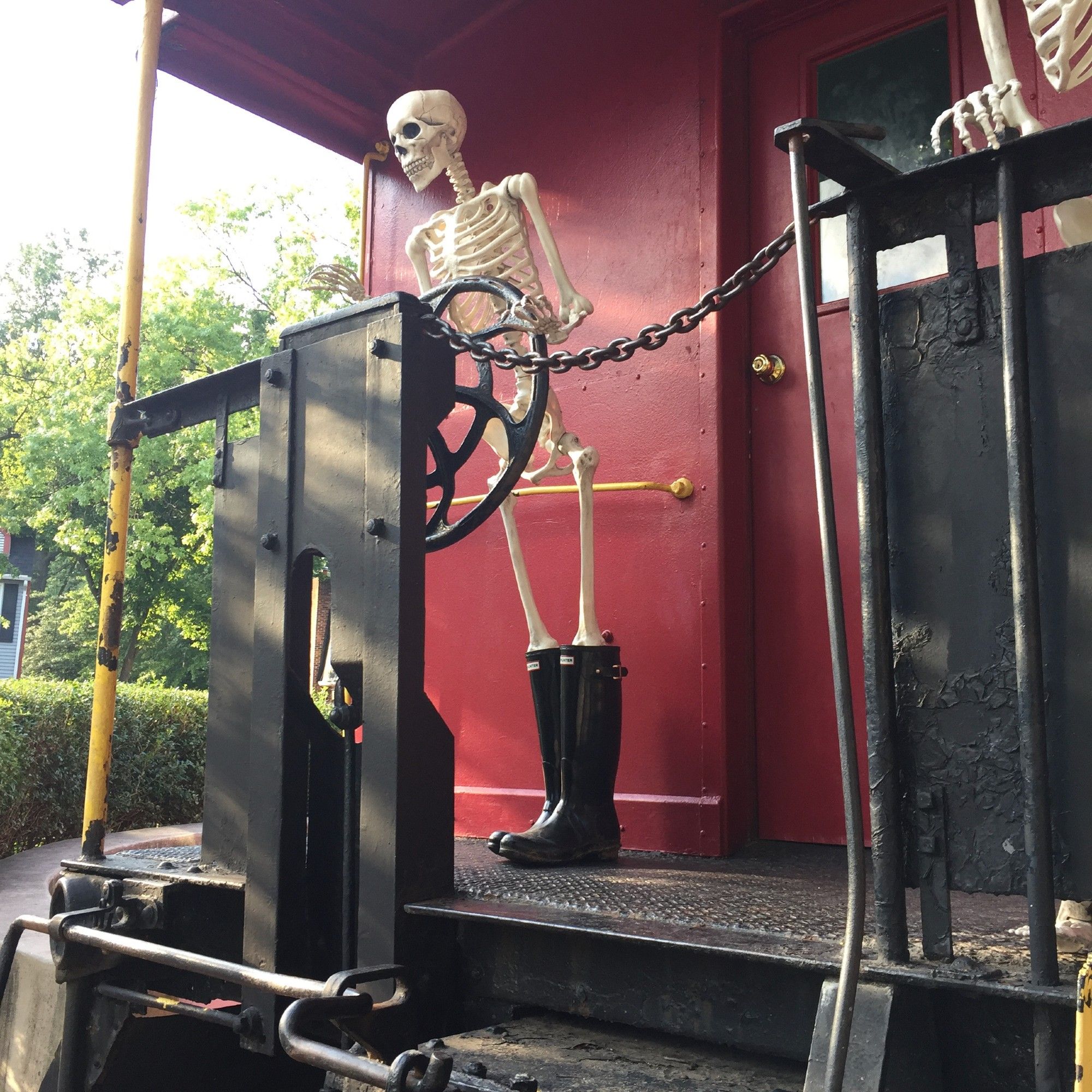 A pair of plastic halloween skeletons "play" on a parked decommissioned red train caboose. They're standing at the back of the car. The one wearing Black hunter boots pretends to steer. The one in the black knit cap hangs on for dear life.