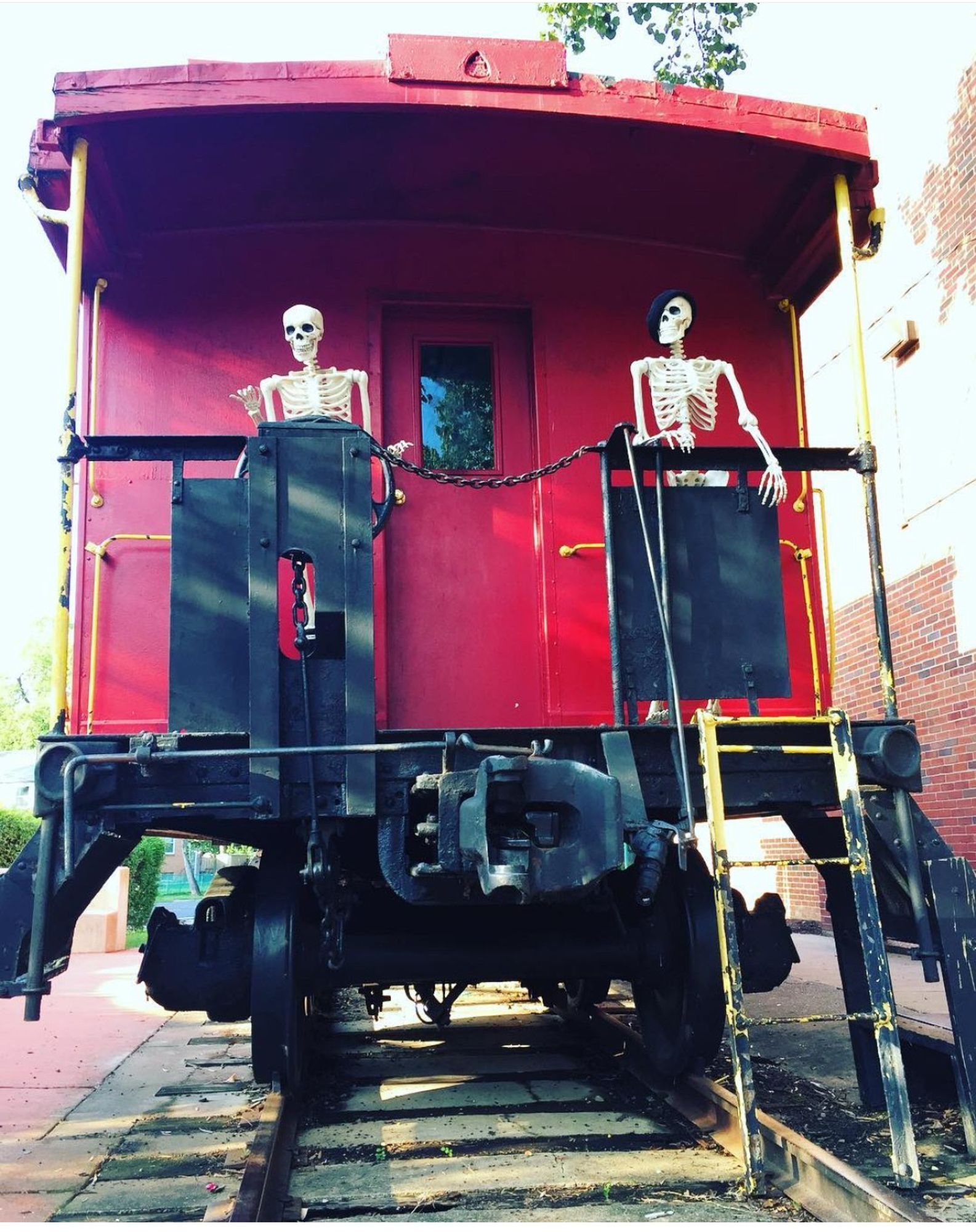 A pair of plastic halloween skeletons "play" on a parked decommissioned red train caboose. They're standing at the back of the car. The one wearing Black hunter boots pretends to steer. The one in the black knit cap hangs on for dear life.