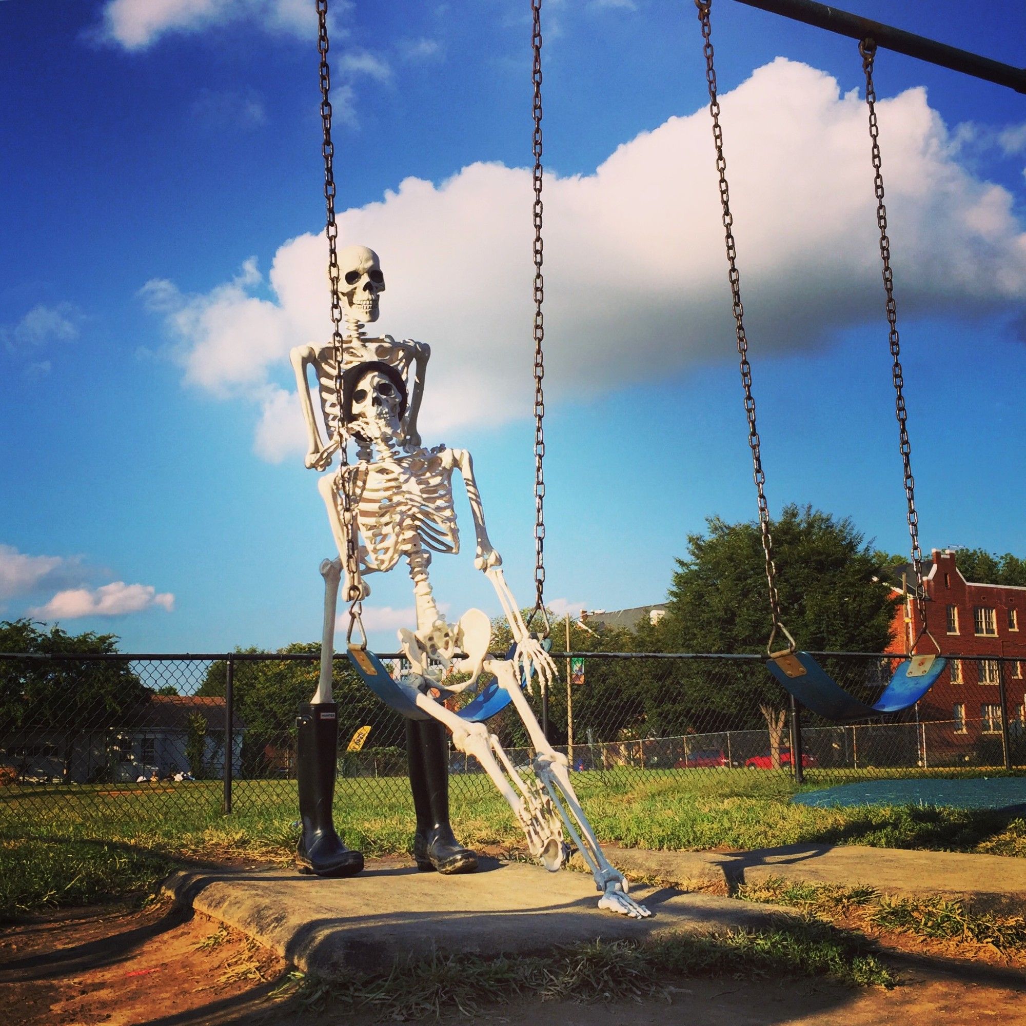 A pair of plastic human halloween skeletons on a playground on a bright and sunny afternoon with blue skies and fluffy white clouds overhead. One skeleton sits in a blue swing, wearing a black knit cap on his head. The other skeleton stands behind the swing in a pair of black kneehigh rainboots.