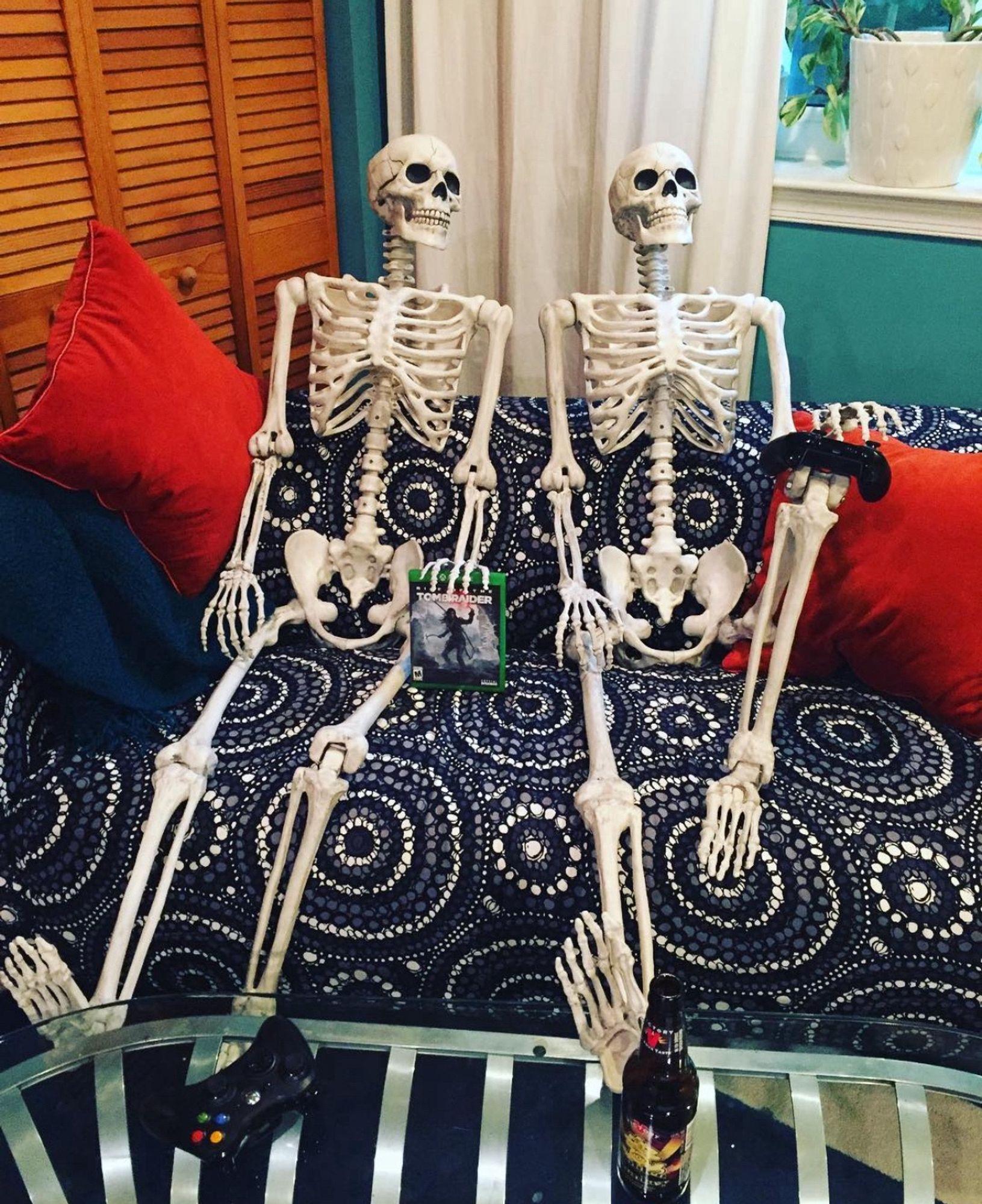 A pair of skeletons sit on an ikea couch with a dramatic black and white textile cover. They have a beer, a game controller and a copy of Tomb Raider.