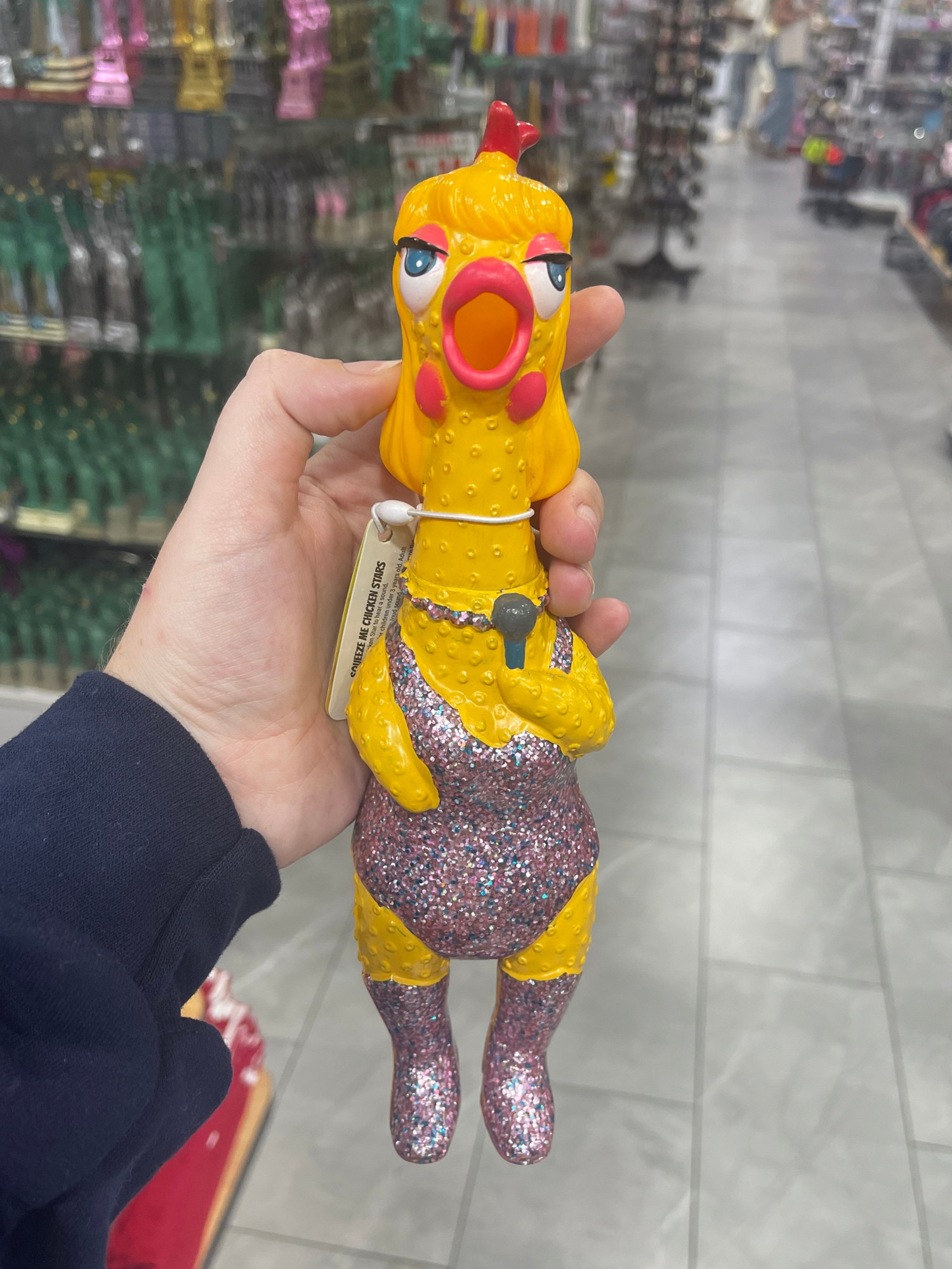 The yellow rubber chicken, wearing a sparkly purple/pink bodysuit and matching boots abd necklace with pink eyeshadow and blue eyes holding a microphone as if it’s taylor in the eras tour — sold at a nyc tourist shop 