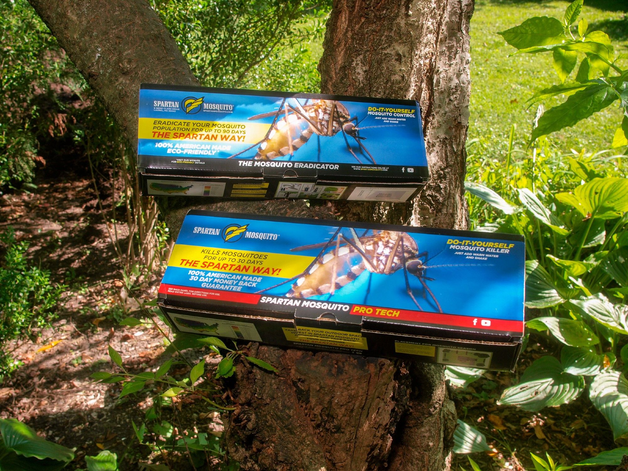 Two long, colorful boxes resting on the trunk of a cherry tree in a sunny yard. One box is labeled, "Spartan Mosquito Eradicator," the other "Spartan Mosquito Pro Tech." Both boxes feature a photograph of a mosquito and are covered with various false claims (e.g., "kills mosquitoes" and "eradicate your mosquito population for up to 90 days").