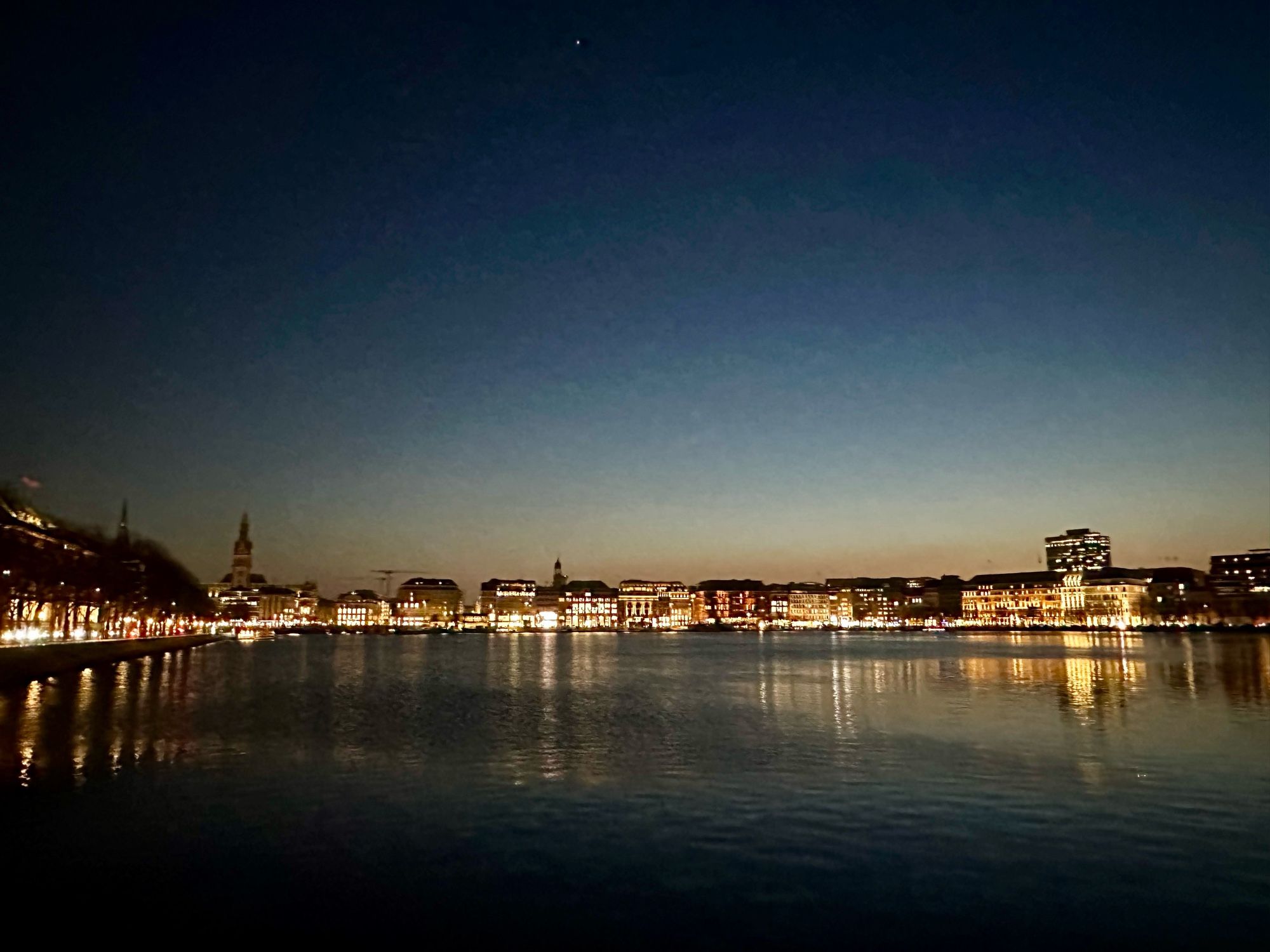 Das Bild zeigt die Hamburger Binnenalster hell erleuchtet im Abendlicht kurz nach Sonnenuntergang. Der Blick richtet sich auf den Jungfernstieg und das Rathaus im Hintergrund. Rechts ist der Neue Jungfernstieg mit dem Hotel Vier Jahreszeiten und links der Ballindamm mit der HapagLloyd-Zentrale und der Europapassage zu sehen.