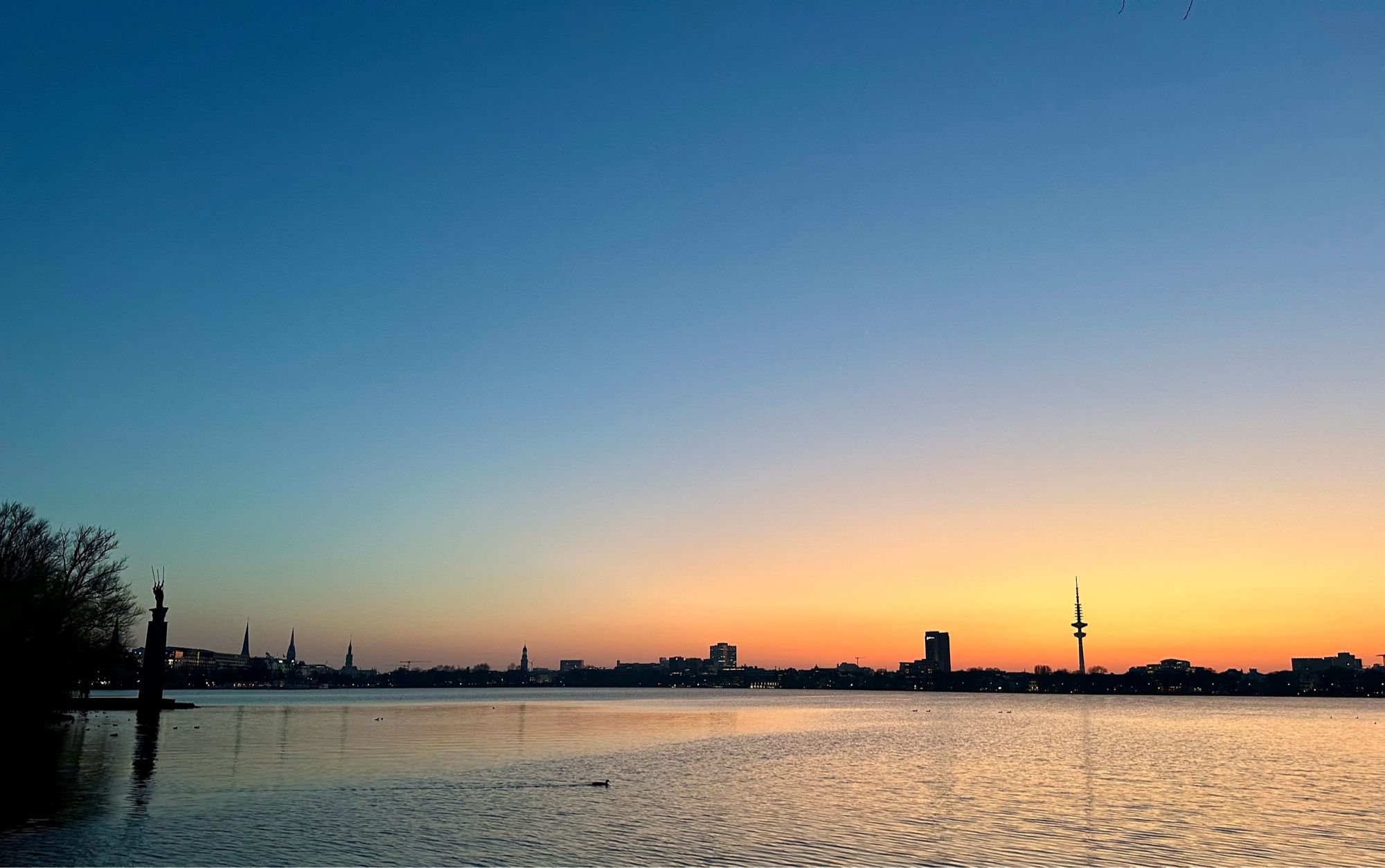 Das Bild zeigt die Außenalster in Hamburg in Blickrichtung Innenstadt im Sonnenuntergang. Im Hintergrund sind diverse Kirchtürme und der Fernsehturm zusehen.