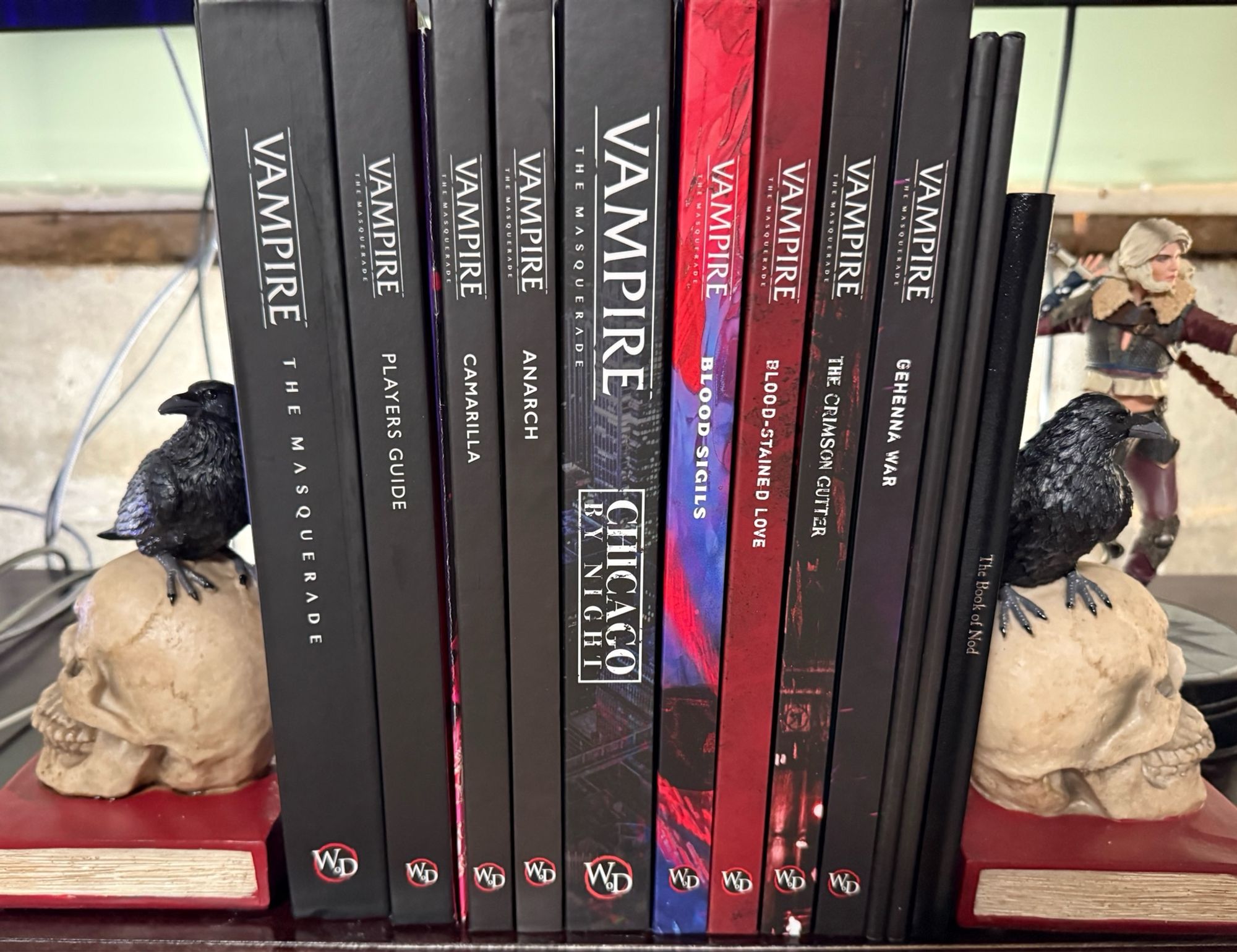 Collection of Vampire the Masquerade 5th edition books held up by bookends statuettes of a crow perched on a human skull that’s resting on a book.