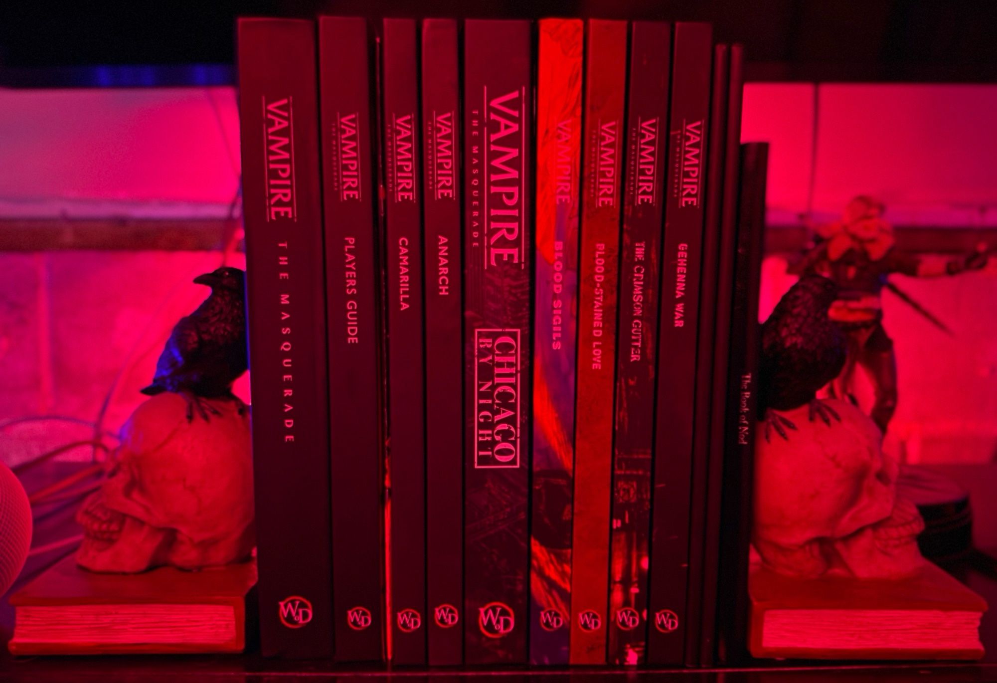 Collection of Vampire the Masquerade 5th edition books held up by bookends statuettes of a crow perched on a human skull that’s resting on a book.