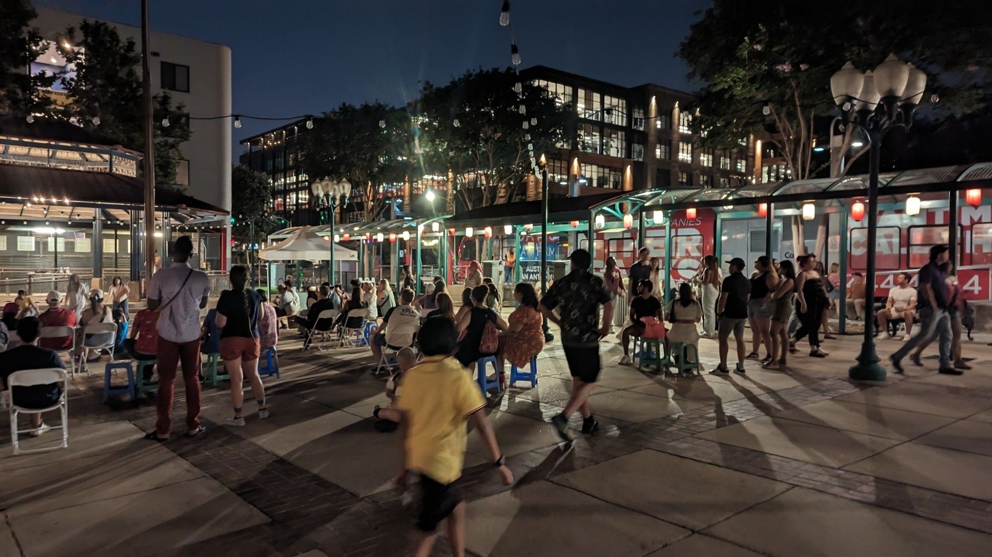 A large concrete tile plaza with a large crowd of people. A child in a yellow shirt is in the foreground. To the left is a stage - to the right is a canopy, behind which is the CapMetro train. Above the CapMetro train, you can see the lights on in the windows of several multi-story buildings. The sky is dark, but with the faintest amount of light, as if the sun set only recently.