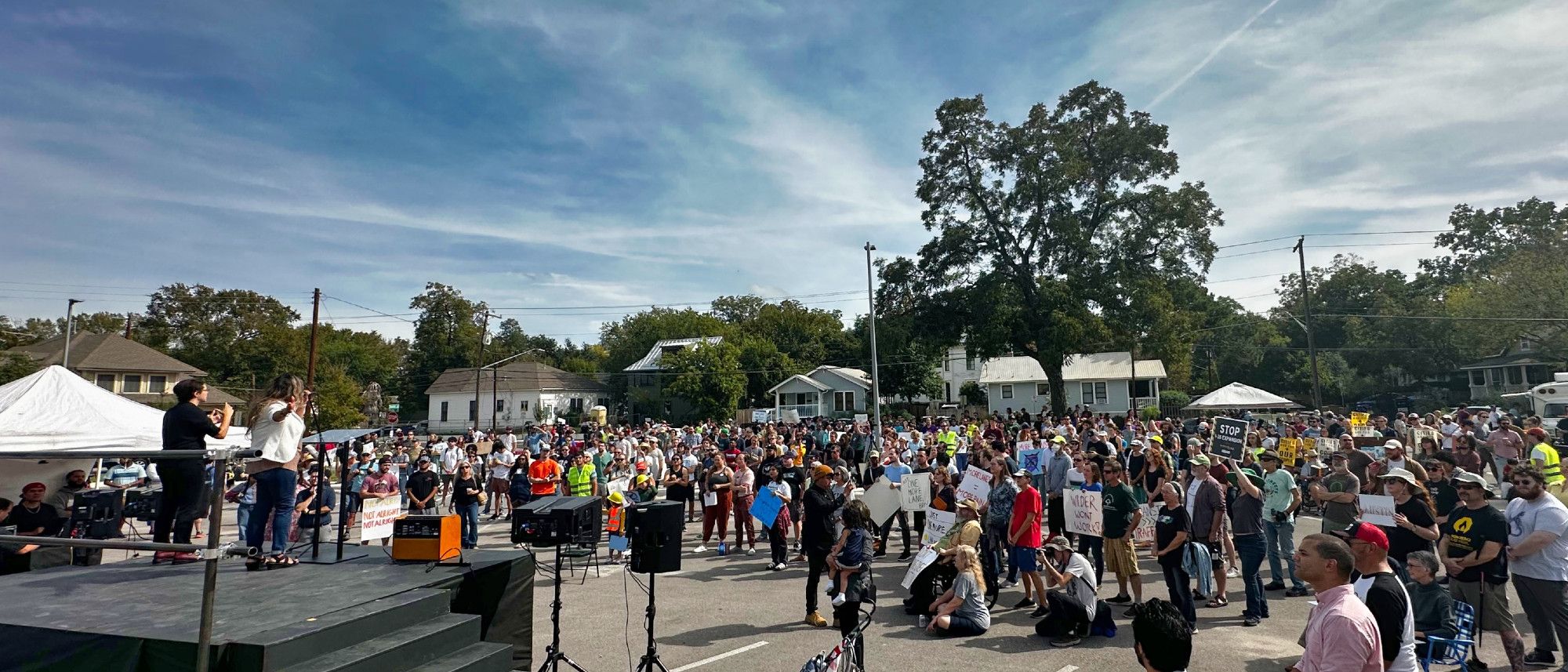 A large crowd (in the hundreds), from all walks of life, gathered outdoors around a stage in an outdoor area. There are small homes visible in the background, and trees stretching up over the crowd. The sky is blue, with moderate cloud cover.