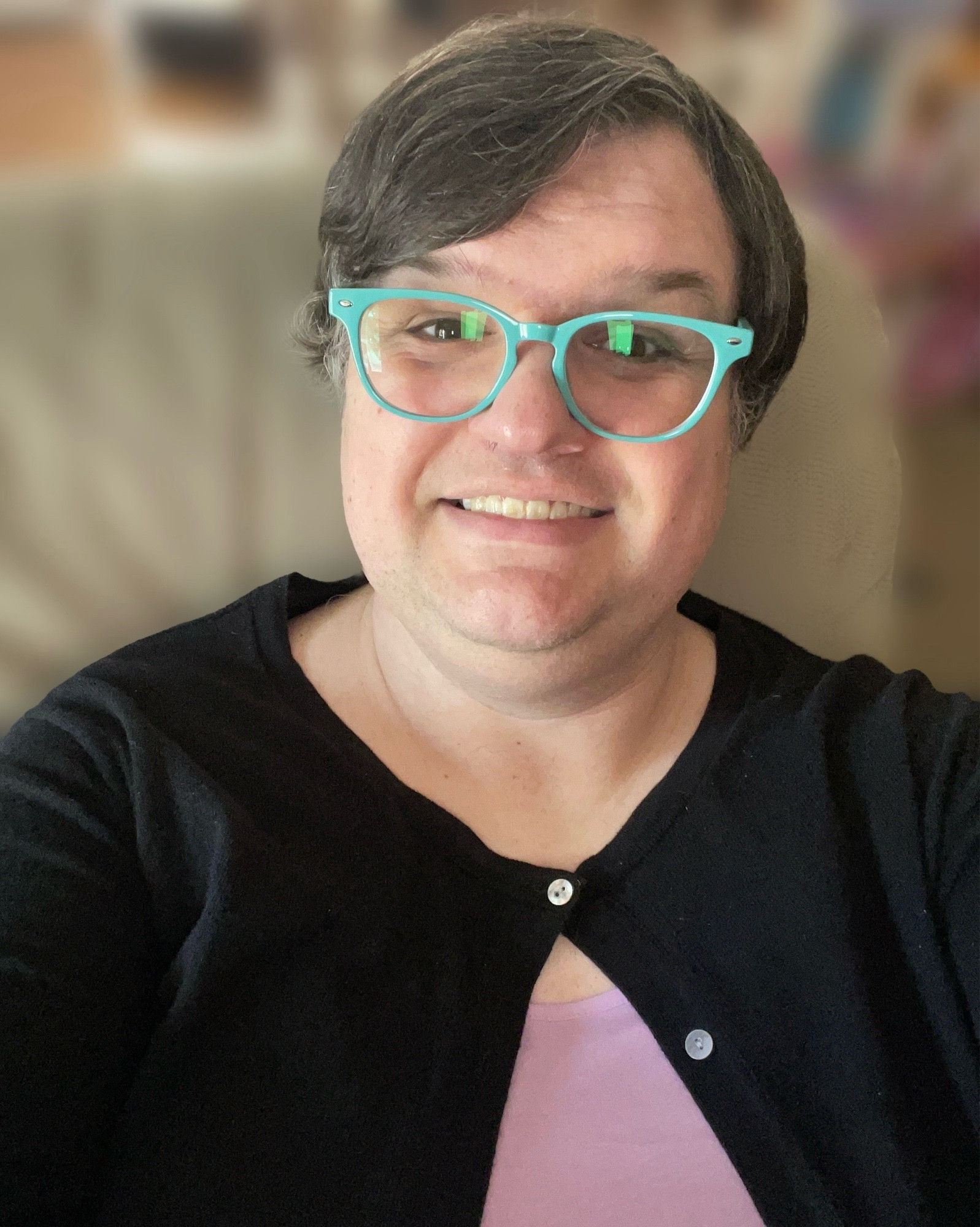 A brown-haired white woman wearing turquoise glasses, a black cardigan and a lavender top is sitting on a tan couch. The background is blurred.