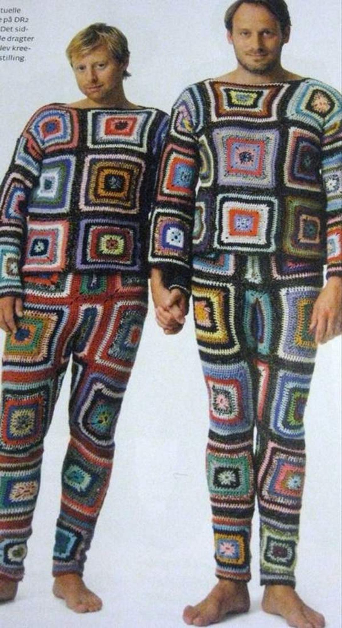 Two men wearing pajamas made entirely of crocheted granny squares. They are barefoot and holding hands.